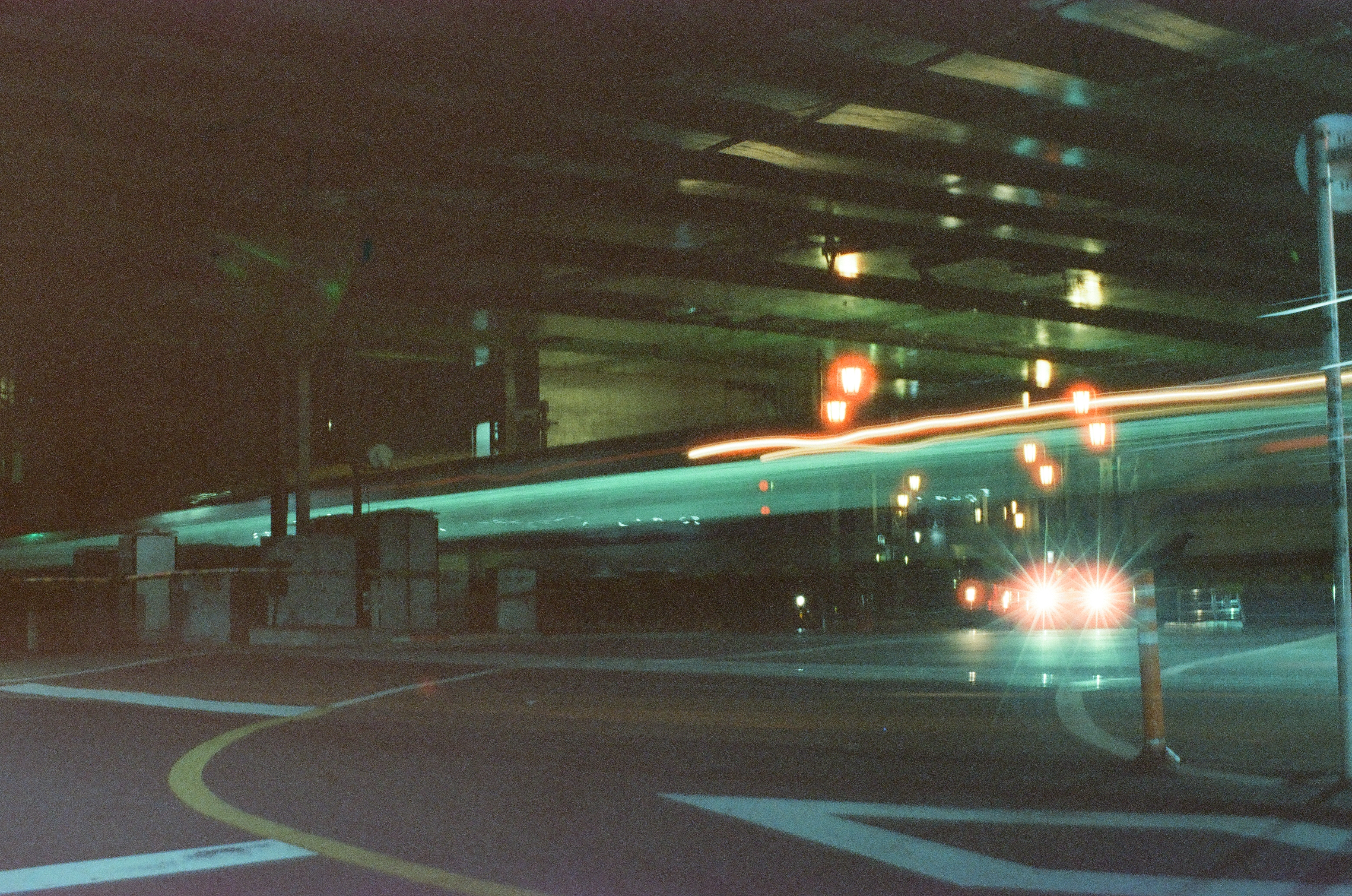 A pale aqua-green light trail stretches across a road beneath a dark elevated overpass, passing rows of red lamps and two bright oncoming headlights.