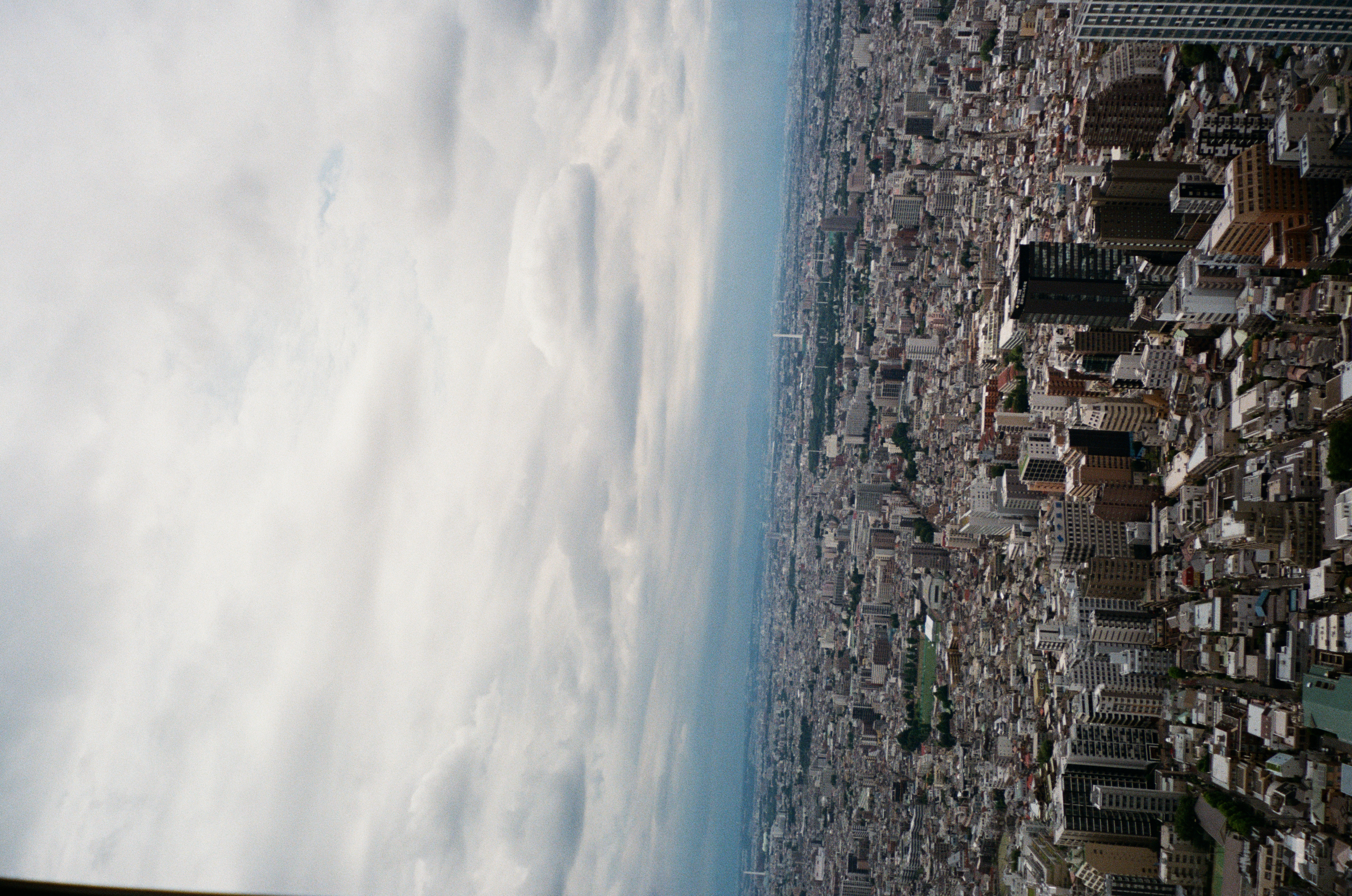 A churning, storm-dark band of cloud presses the horizon above a dense city and a green athletic field stretching into the distance.