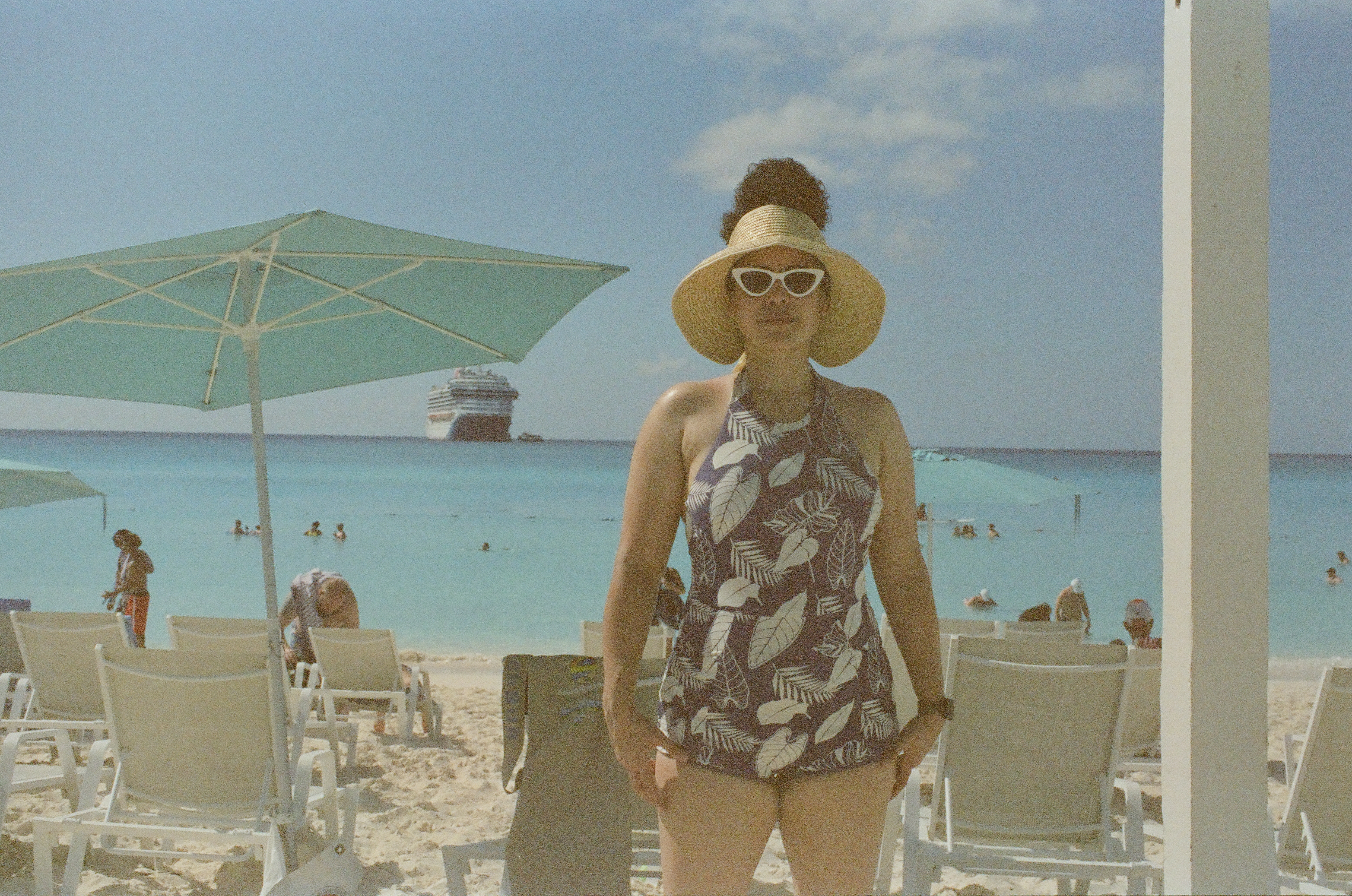 An adult in a hat, white sunglasses, and a patterned swimsuit stands among chairs; swimmers and a ship appear behind.