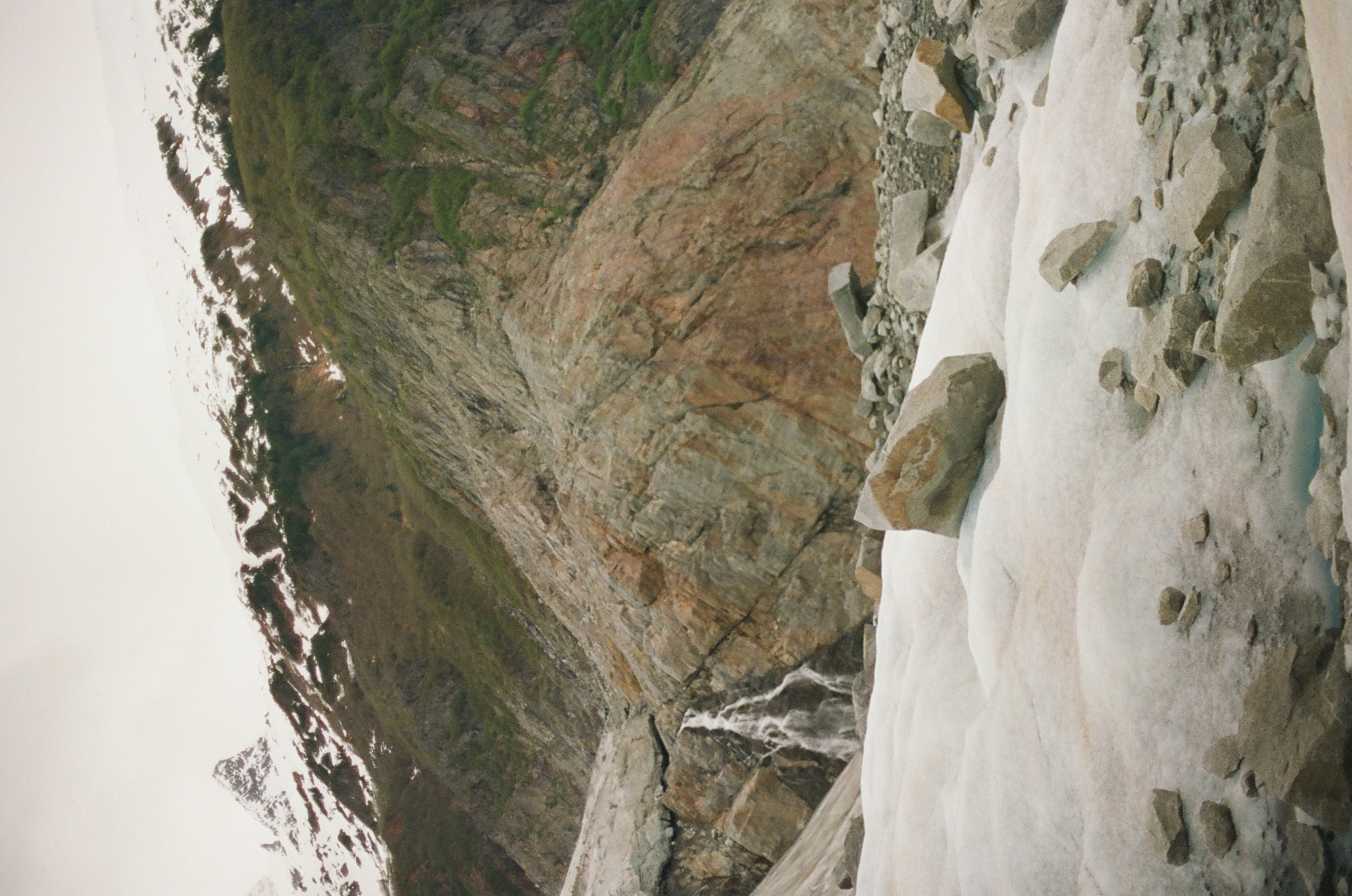 Thin waterfalls descend a rust-colored cliff behind a snowbank scattered with rounded gray stones.
