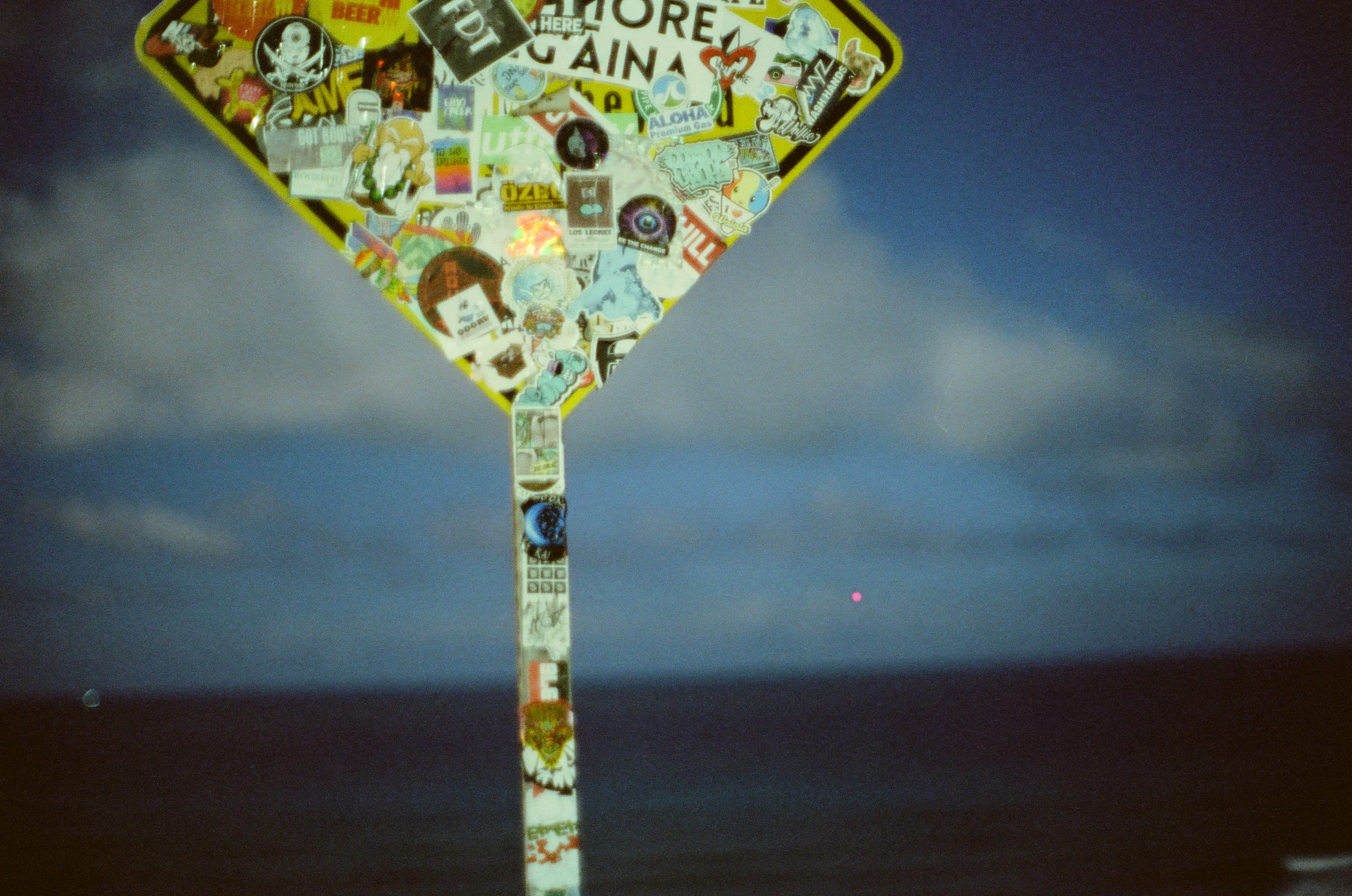 A diamond road sign covered in stickers rises against deep blue clouds with a small reddish moon above.