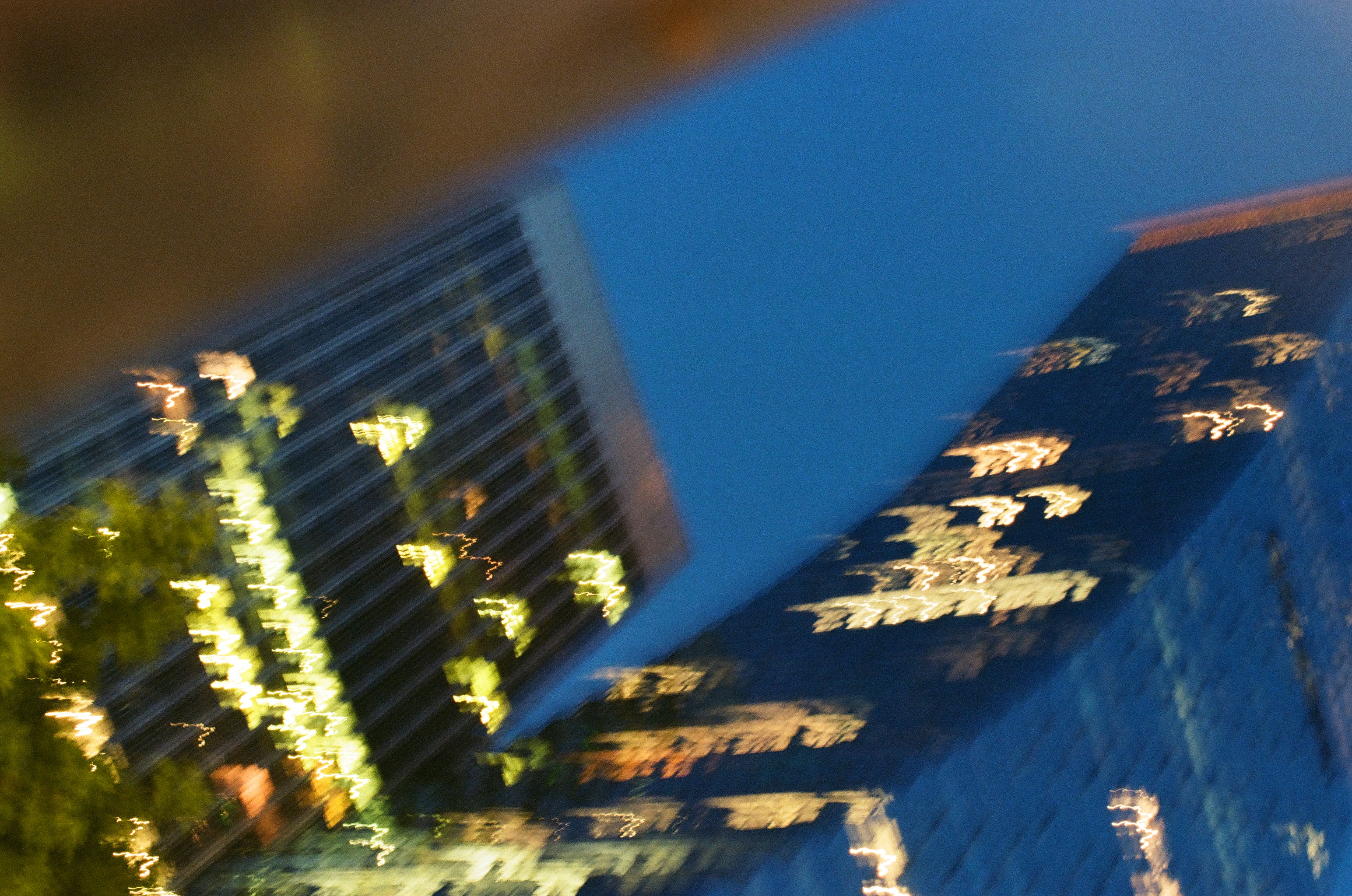 Lit high-rises tilt into blue evening as camera motion smears windows and nearby foliage.