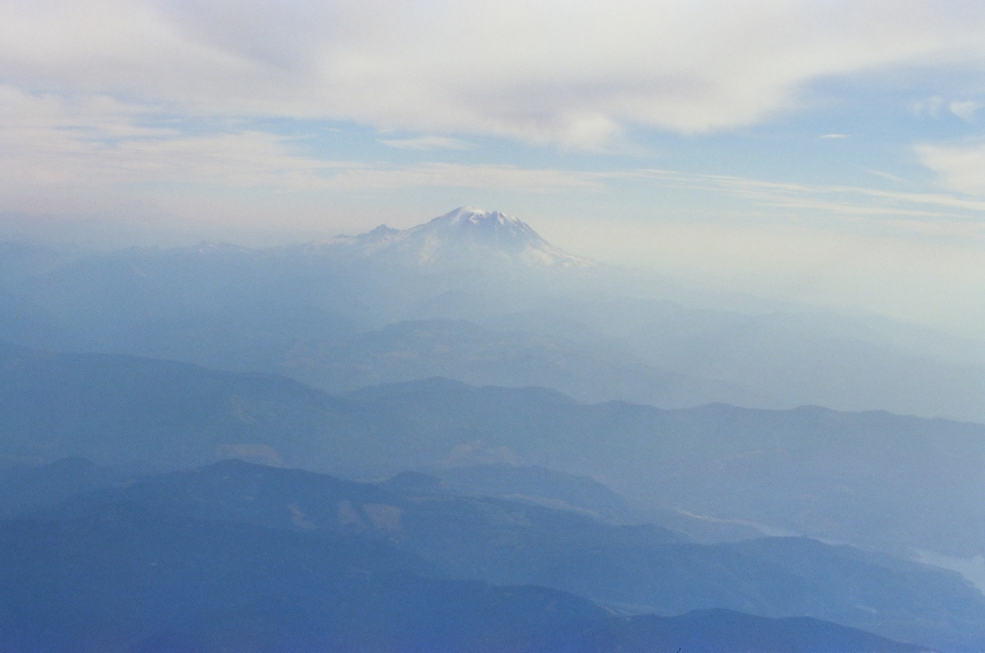 A snow-capped peak sits above hazy blue ridgelines, with a thin pale diagonal streak visible in the sky to its left.