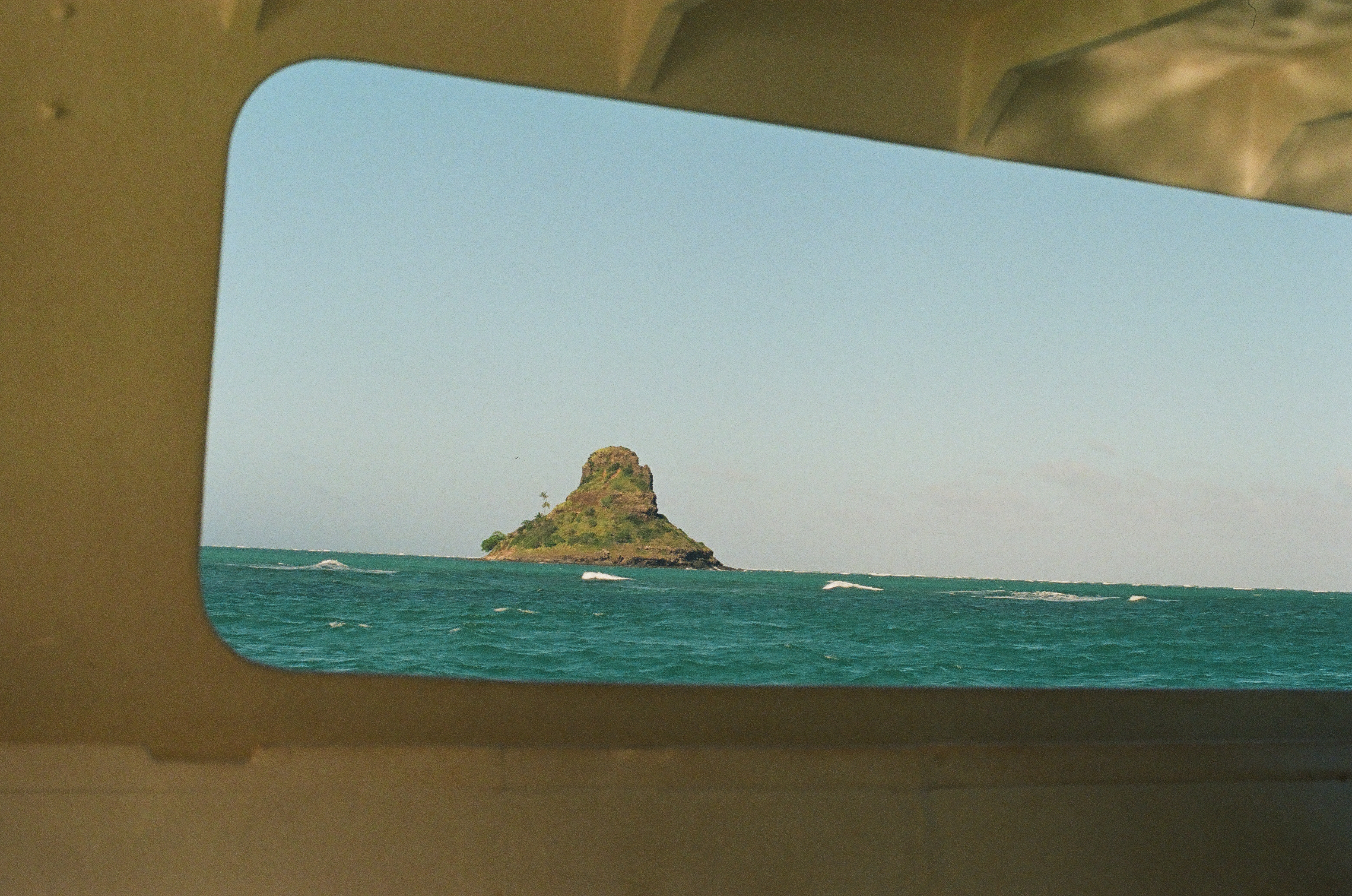 Through the same boat window, a small conical islet topped with palms sits on the horizon above choppy turquoise water streaked with whitecaps.