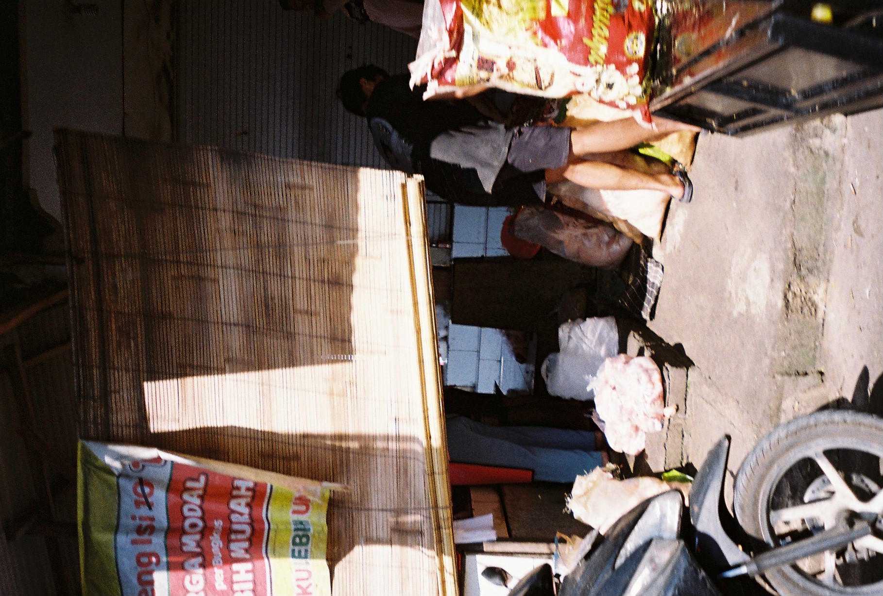 A person works among bags inside a dark roadside stall as scooters and bright signs crowd the entrance.