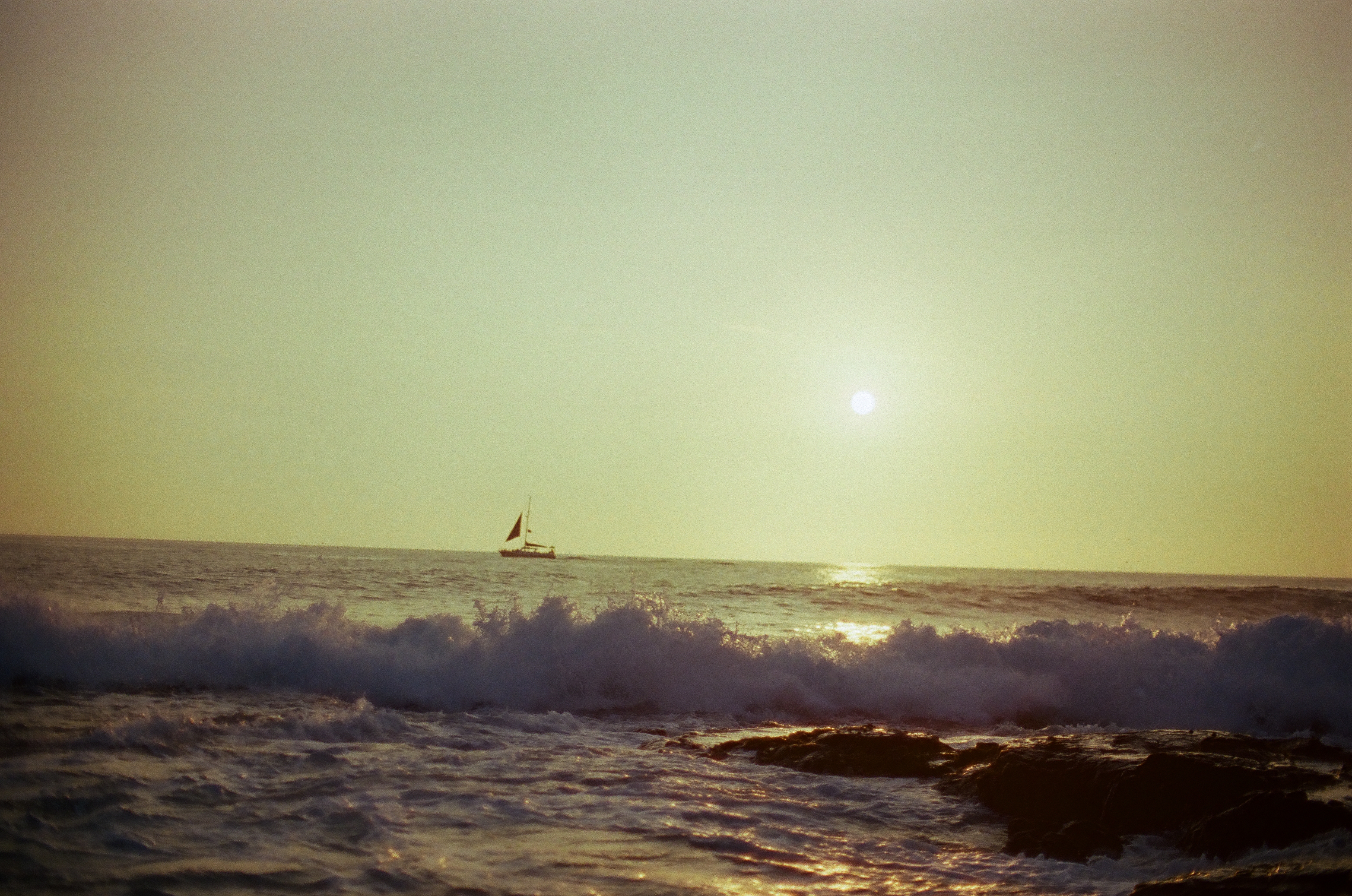 A small sailboat crosses the bright horizon while a dark blue wave breaks over foreground rocks.