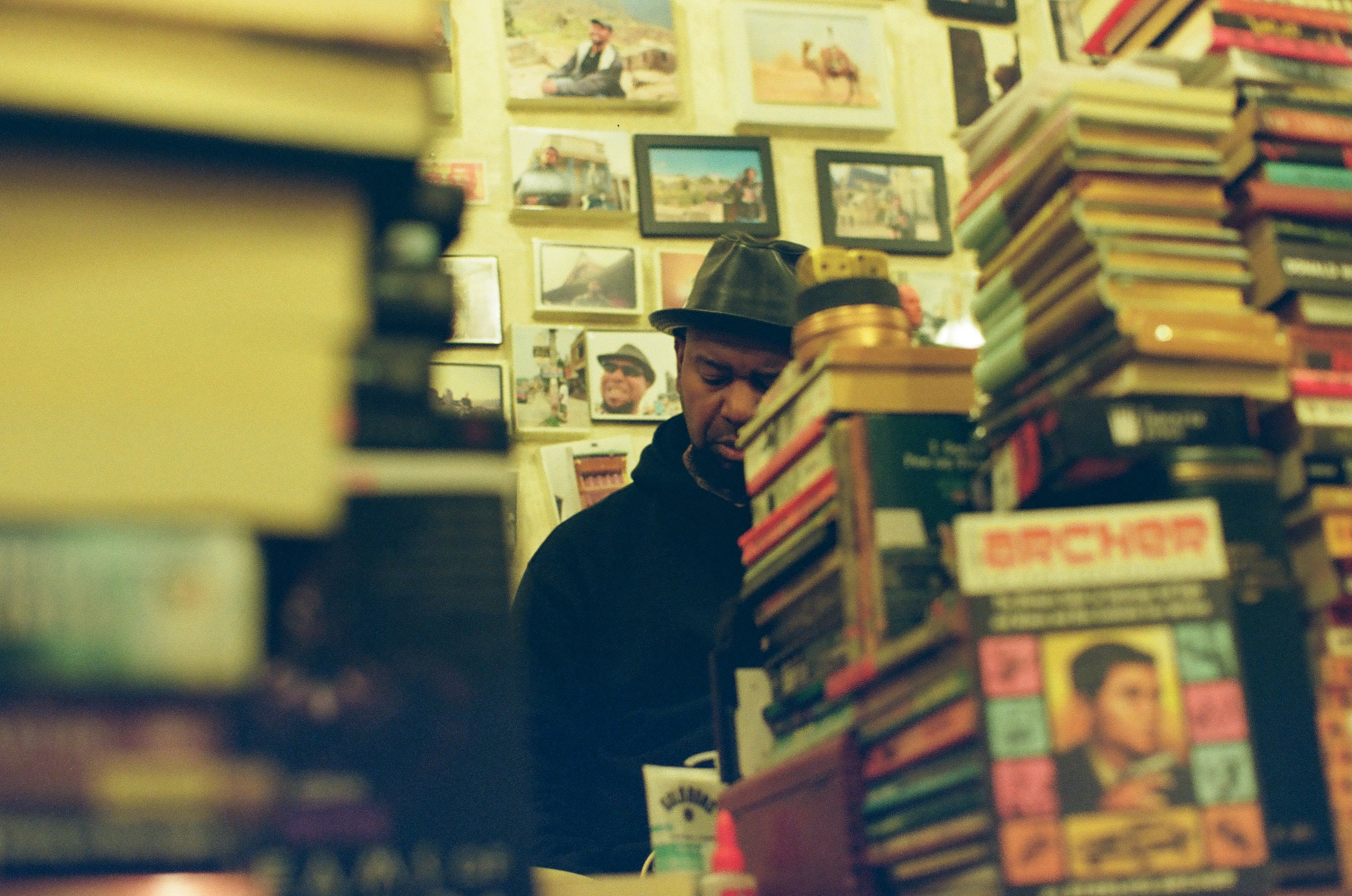 A man in a dark hat looks downward between tall book stacks and a wall of framed pictures.