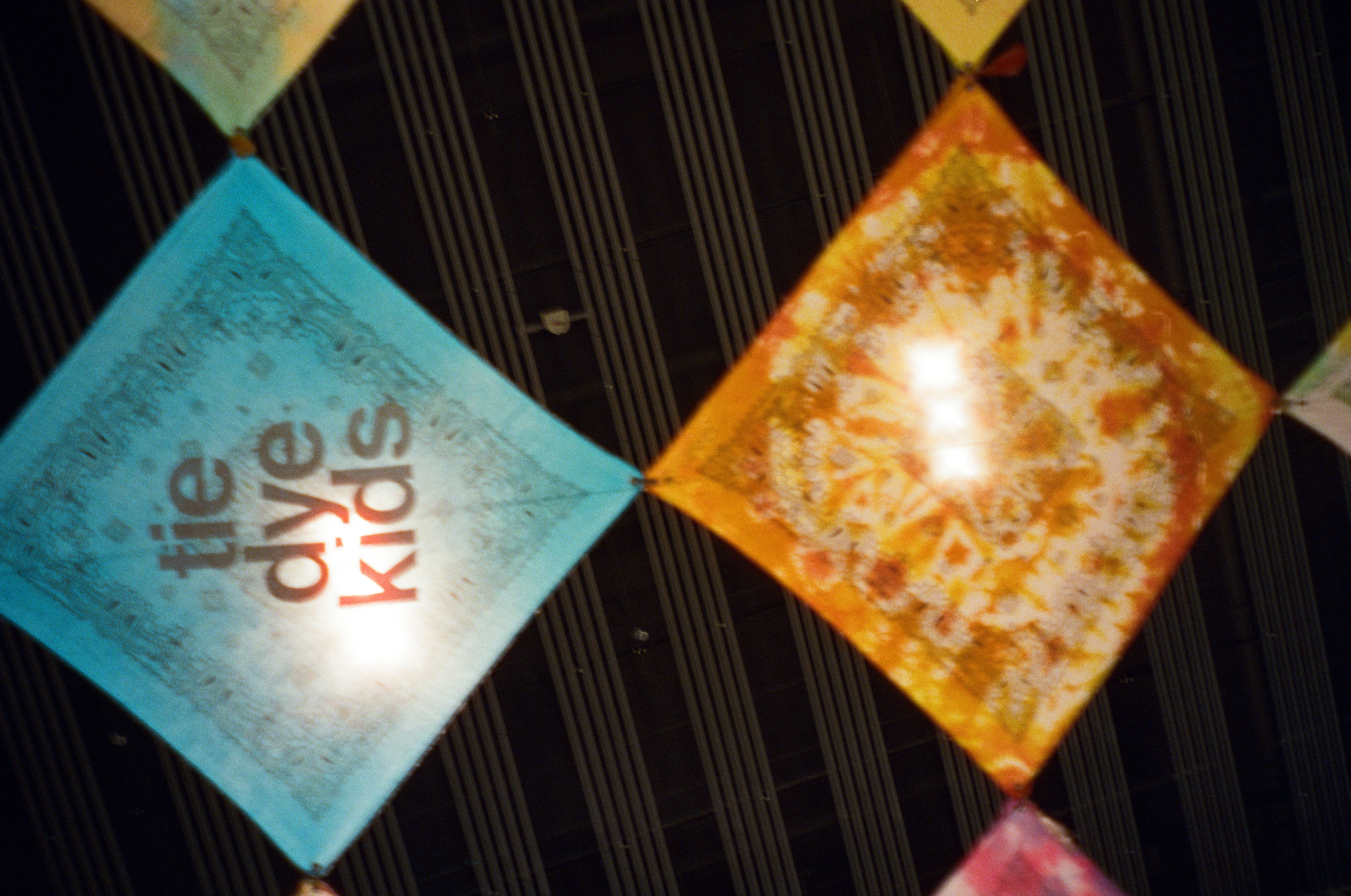 A turquoise fabric panel printed with the words 'tie dye kids' hangs diamond-wise above an orange panel with a sunburst pattern.