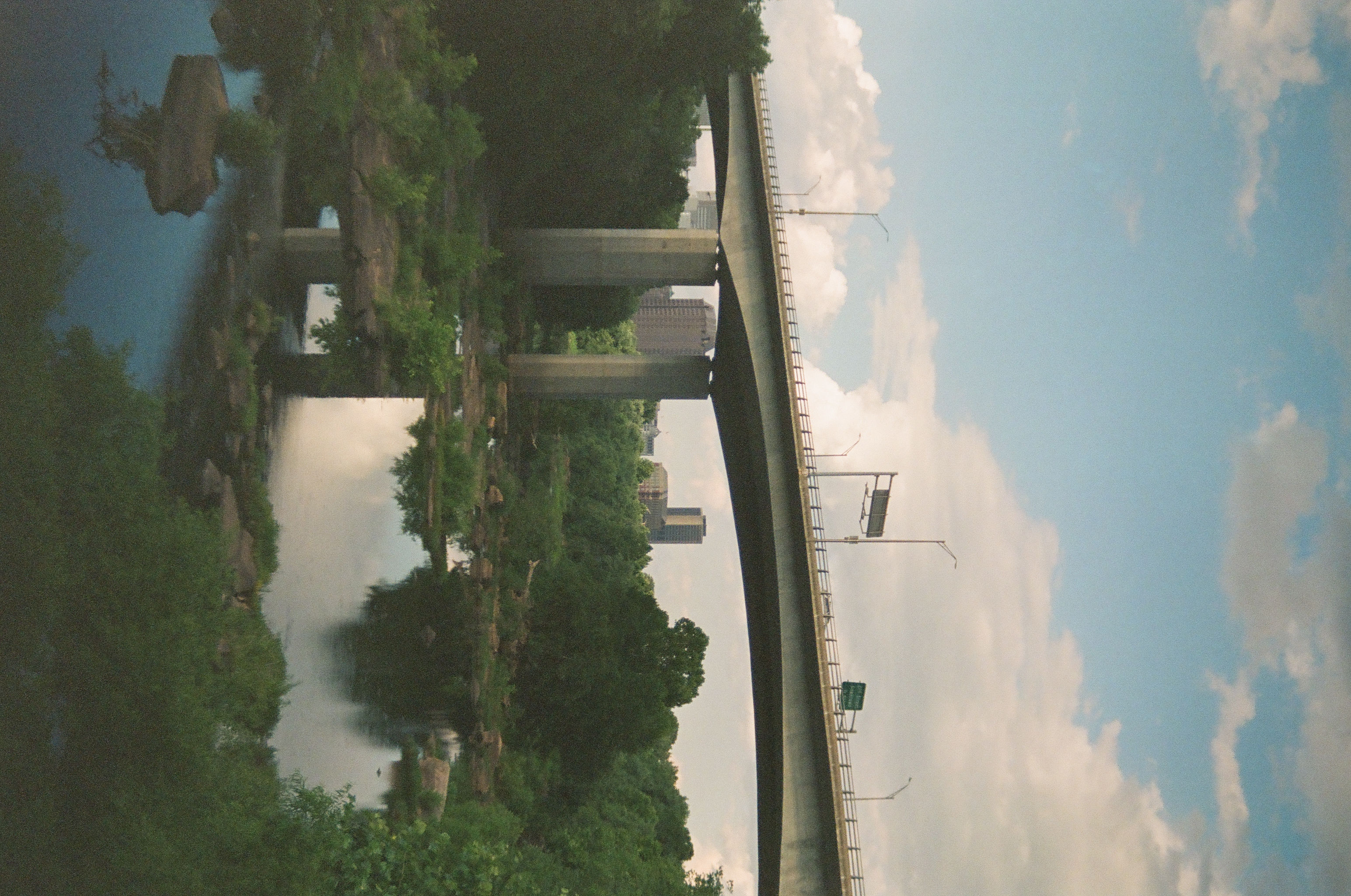 A concrete highway bridge arcs over a river with a few distant buildings visible beneath its deck, mirrored in still water below cumulus clouds.