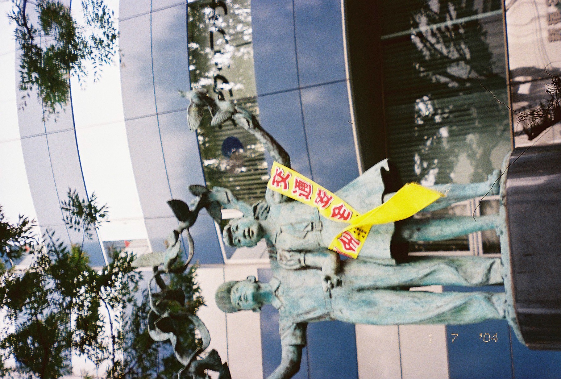 A verdigris bronze statue of two figures releasing doves wears a yellow sash printed with the Japanese characters '交通安全' across its front.