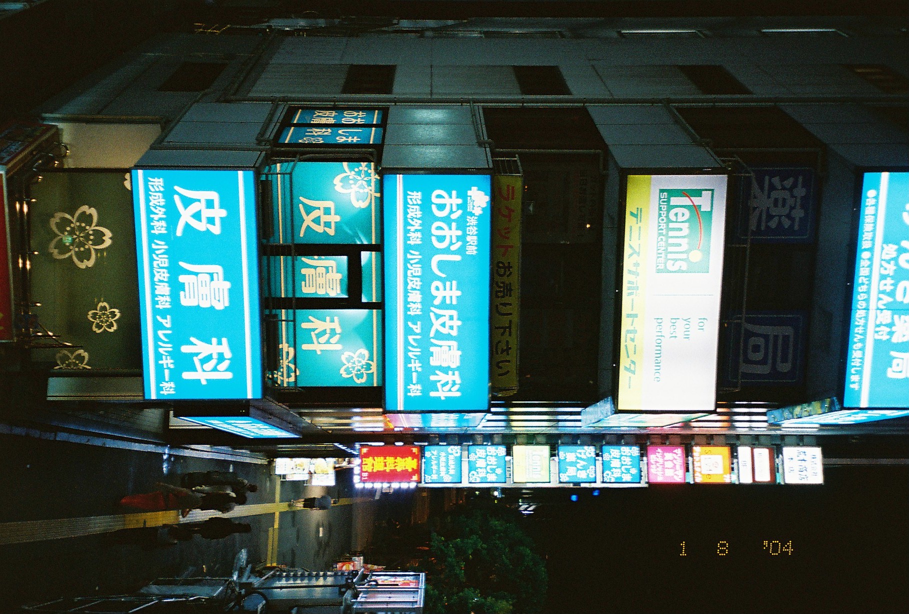 Stacked backlit signs climb a narrow tower at night, including teal panels reading 渋谷駅前 おおしま皮膚科 above repeated 皮膚科 lettering.