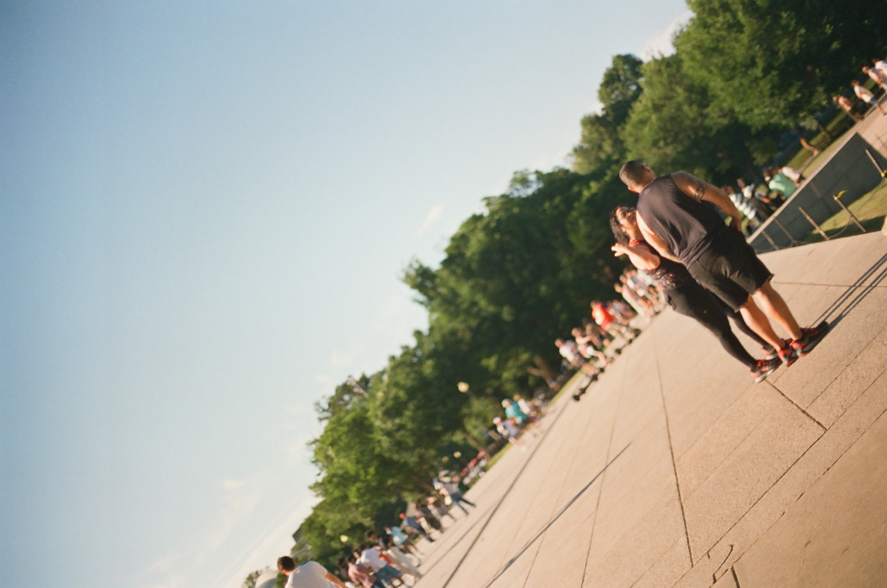 Two people stand close together on a wide paved plaza seen at a steep tilt, with scattered visitors and a line of green trees behind them.