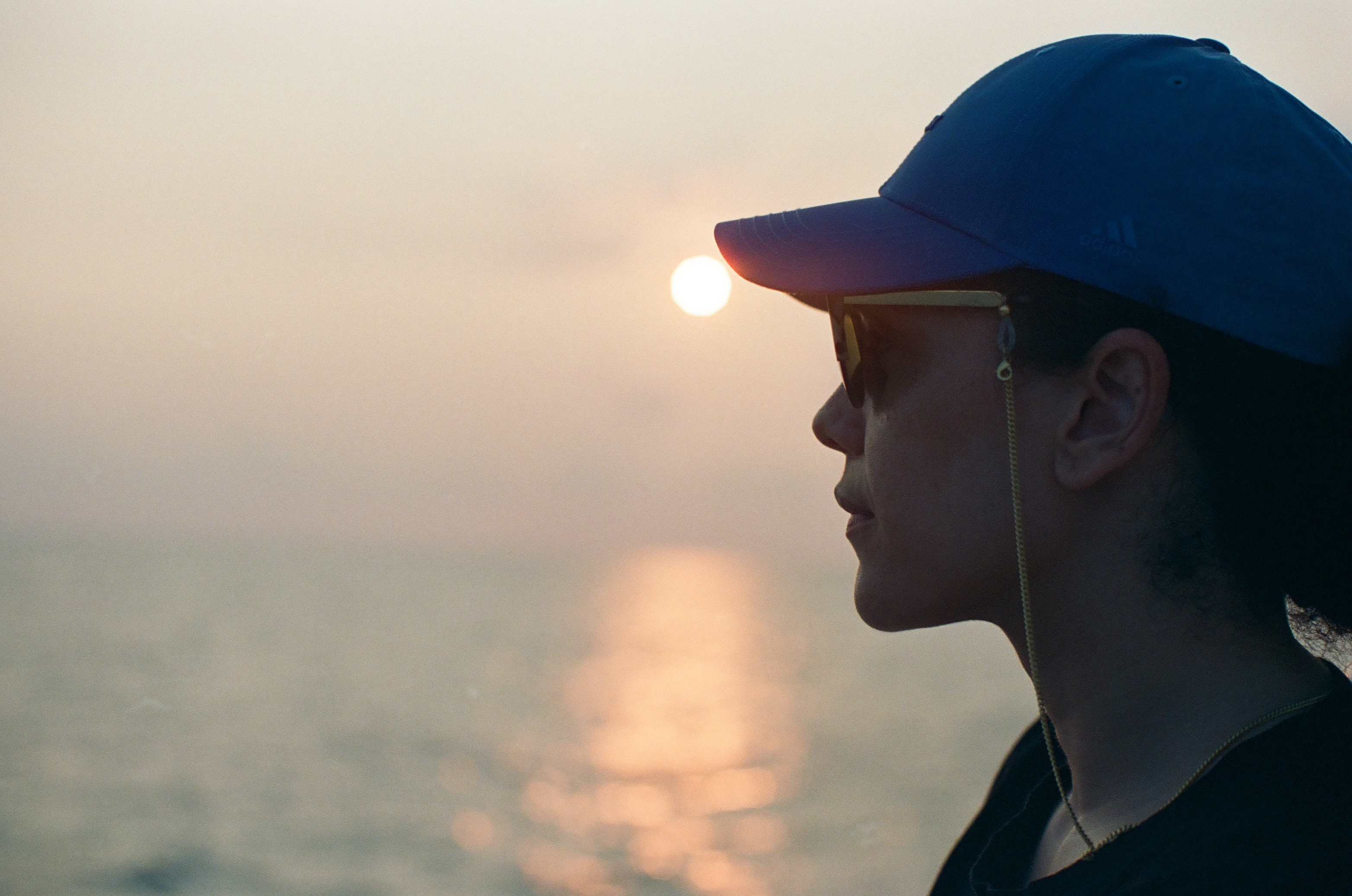 A person in a blue cap and dark glasses appears in profile with the low sun glowing above calm water.