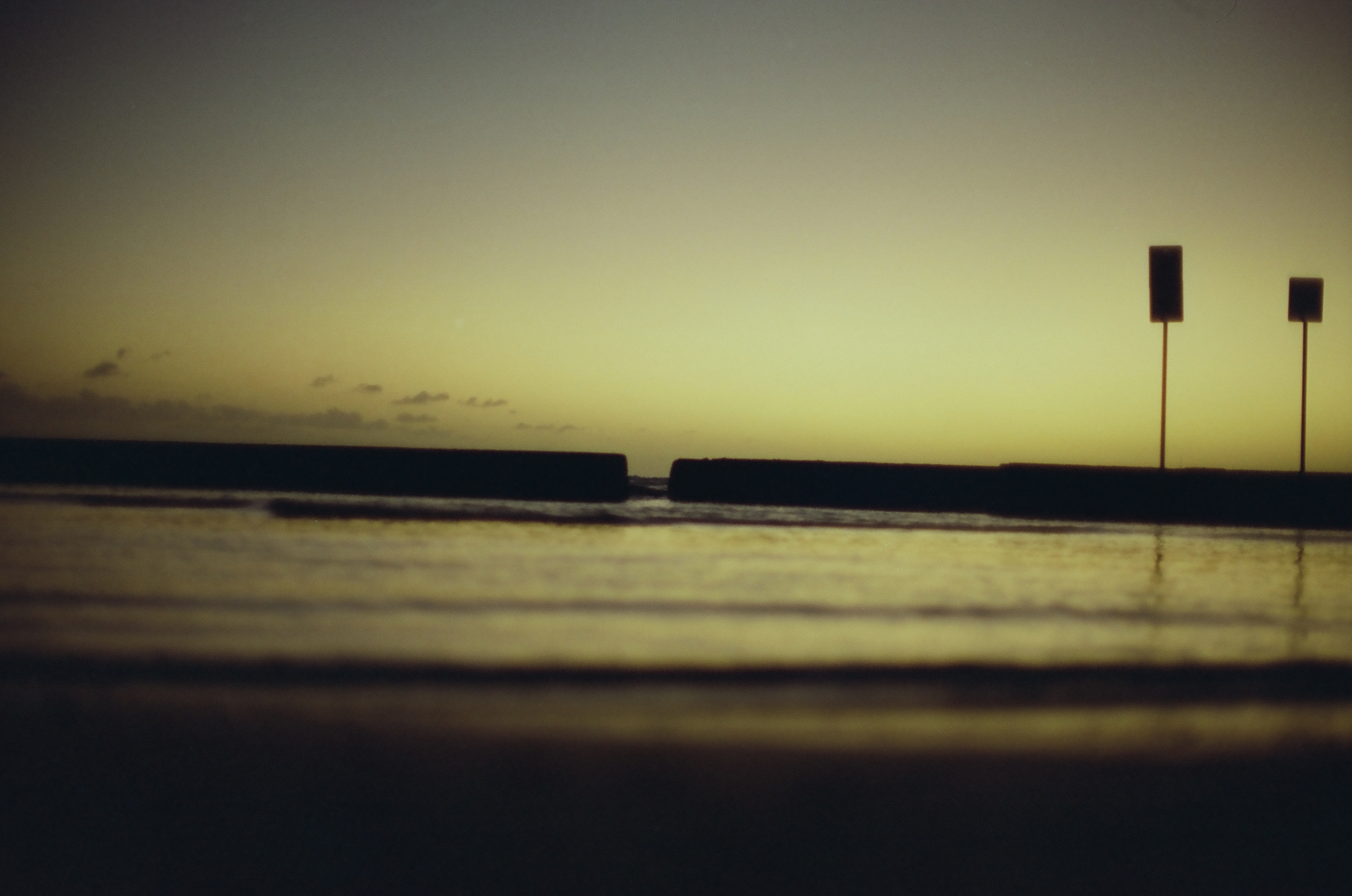 Two narrow signs stand beyond a dark seawall beneath a pale green and gold evening sky.