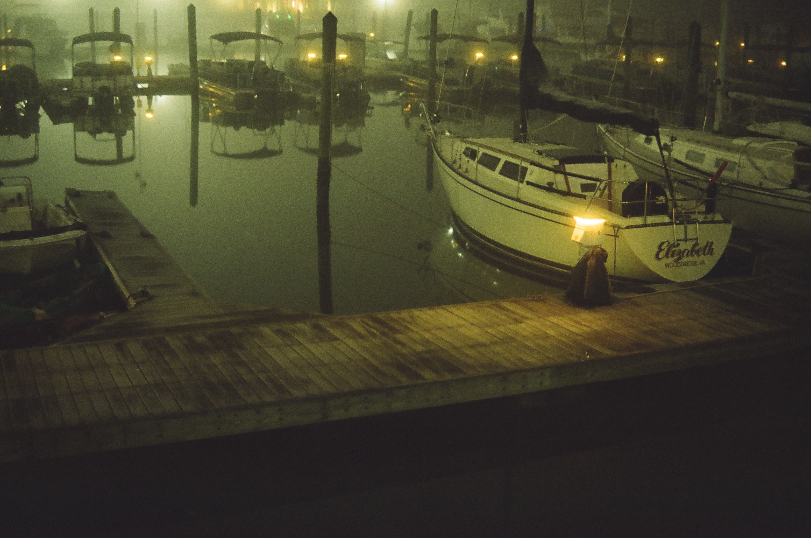 Seen from above, a wooden dock in dense fog holds a sailboat named Elizabeth beside rows of moored boats fading into haze.