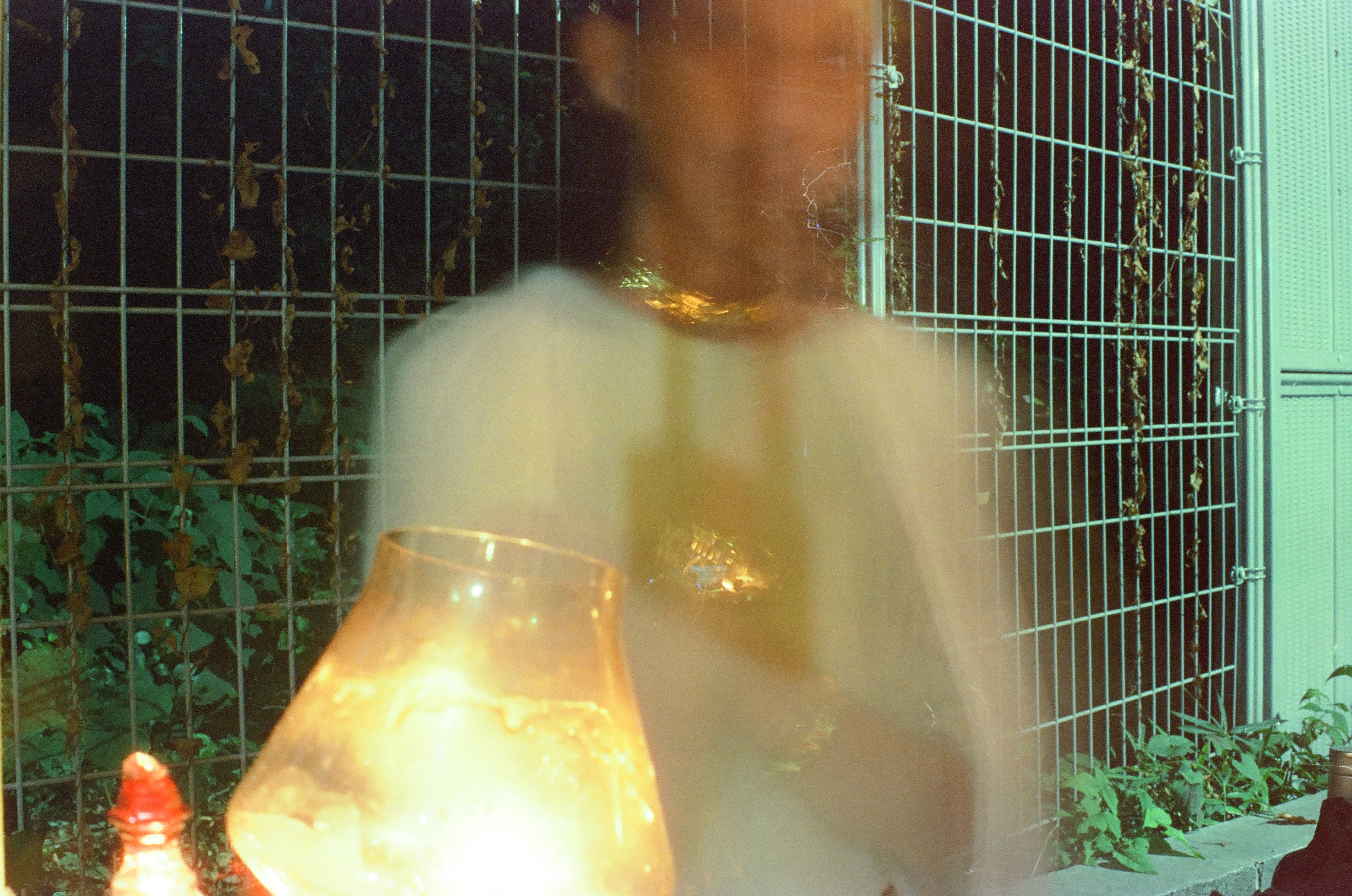 A person in a pale, loose shirt is rendered almost transparent by motion blur behind a glowing amber glass vessel and a vine-covered wire fence panel.