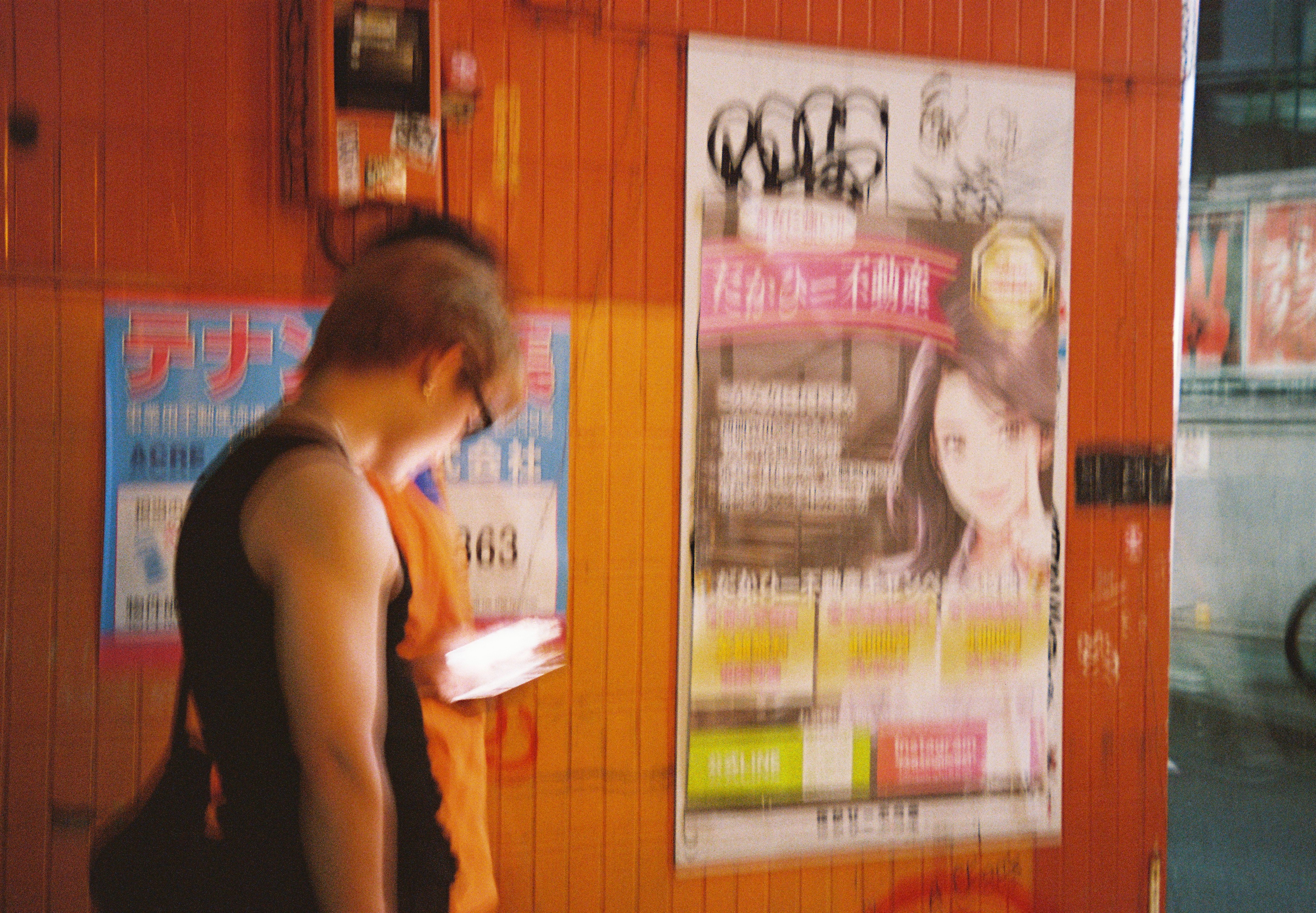 A man in a black tank top looks down at a lit object in his hands beside an orange wall covered in posters.