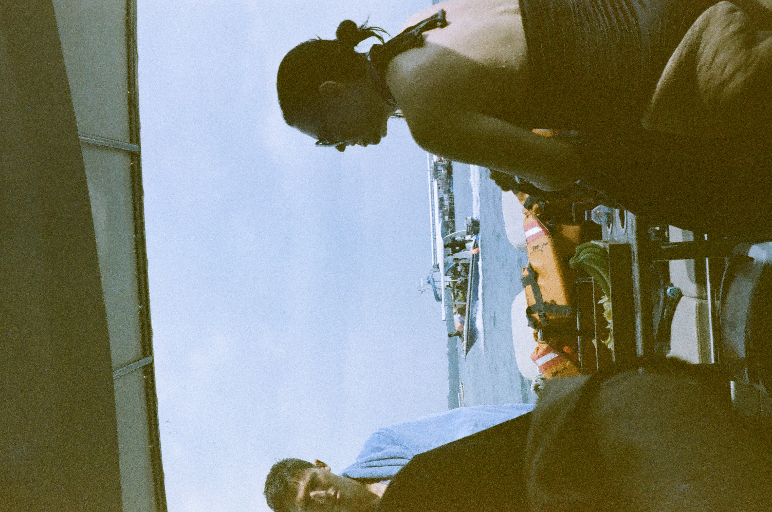 Several seated passengers gather beneath a canopy while bright water and another boat remain visible behind them.