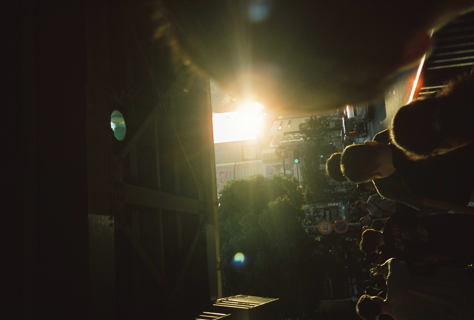 A crowd is reduced to silhouettes beneath dark riveted girders as a low sun flares through the far end of a footbridge.