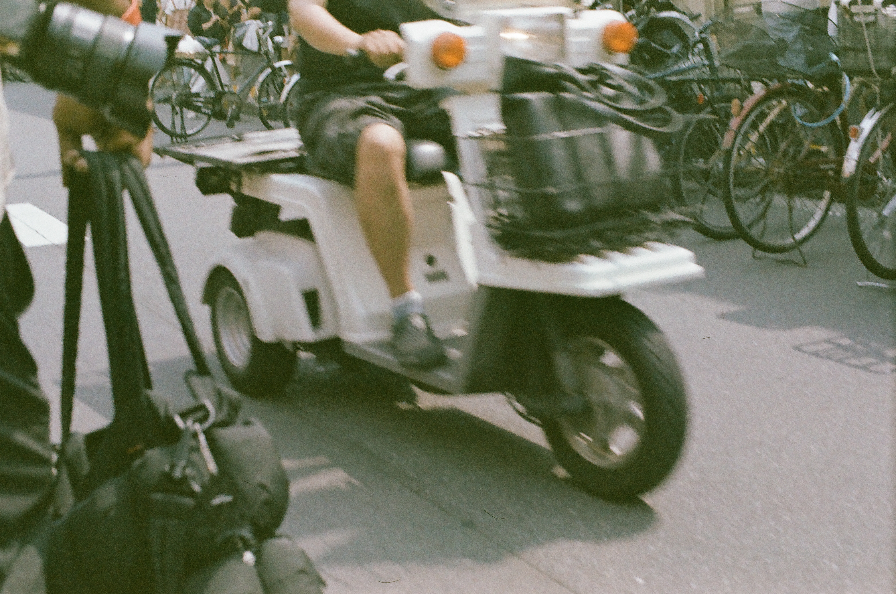 A white three-wheeled scooter with a rider in shorts blurs past a dense row of parked bicycles on a paved street.