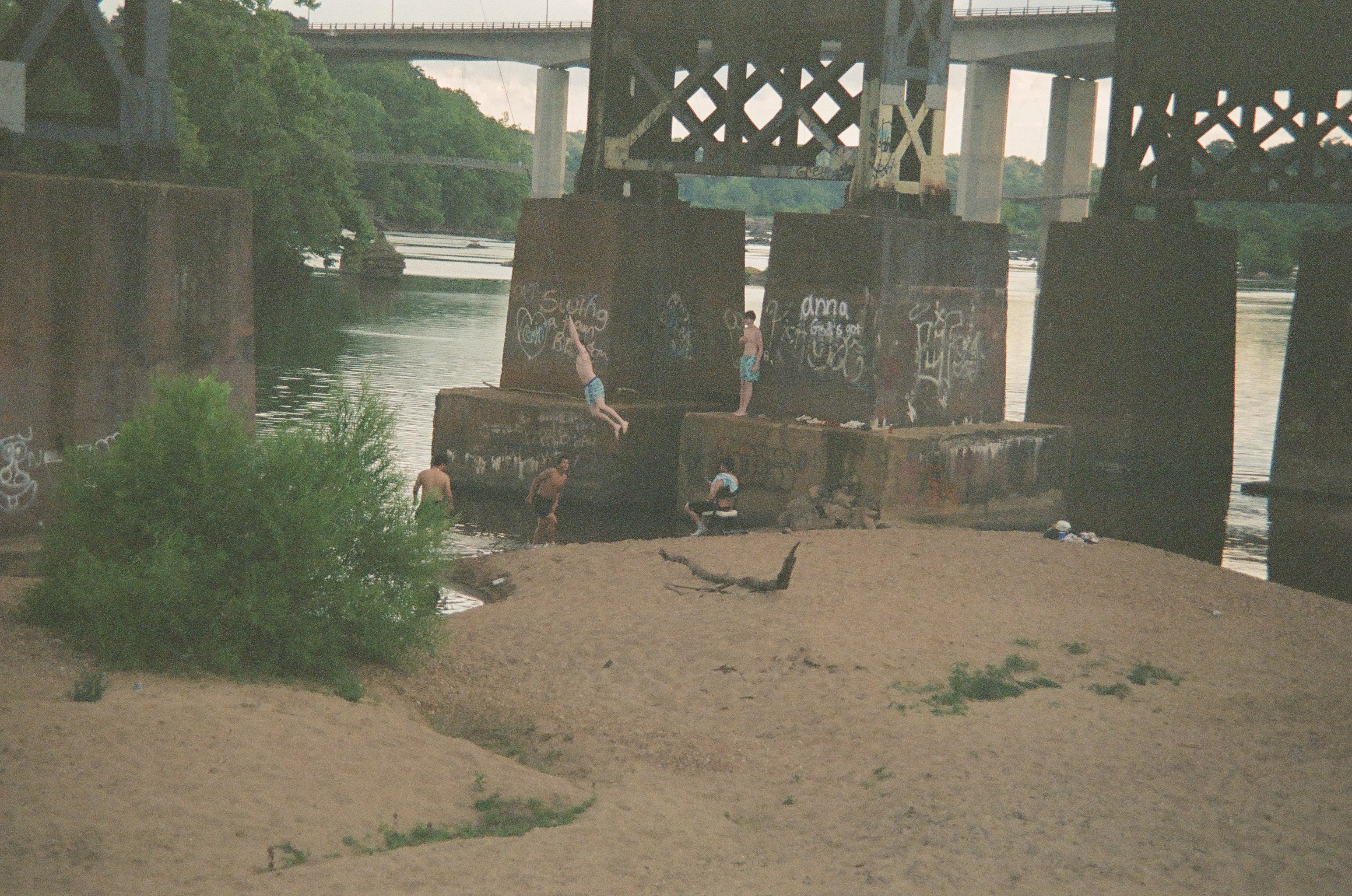 People swim and jump from graffiti-covered concrete piers into a river beside a sandy beach, with a highway bridge visible behind them.