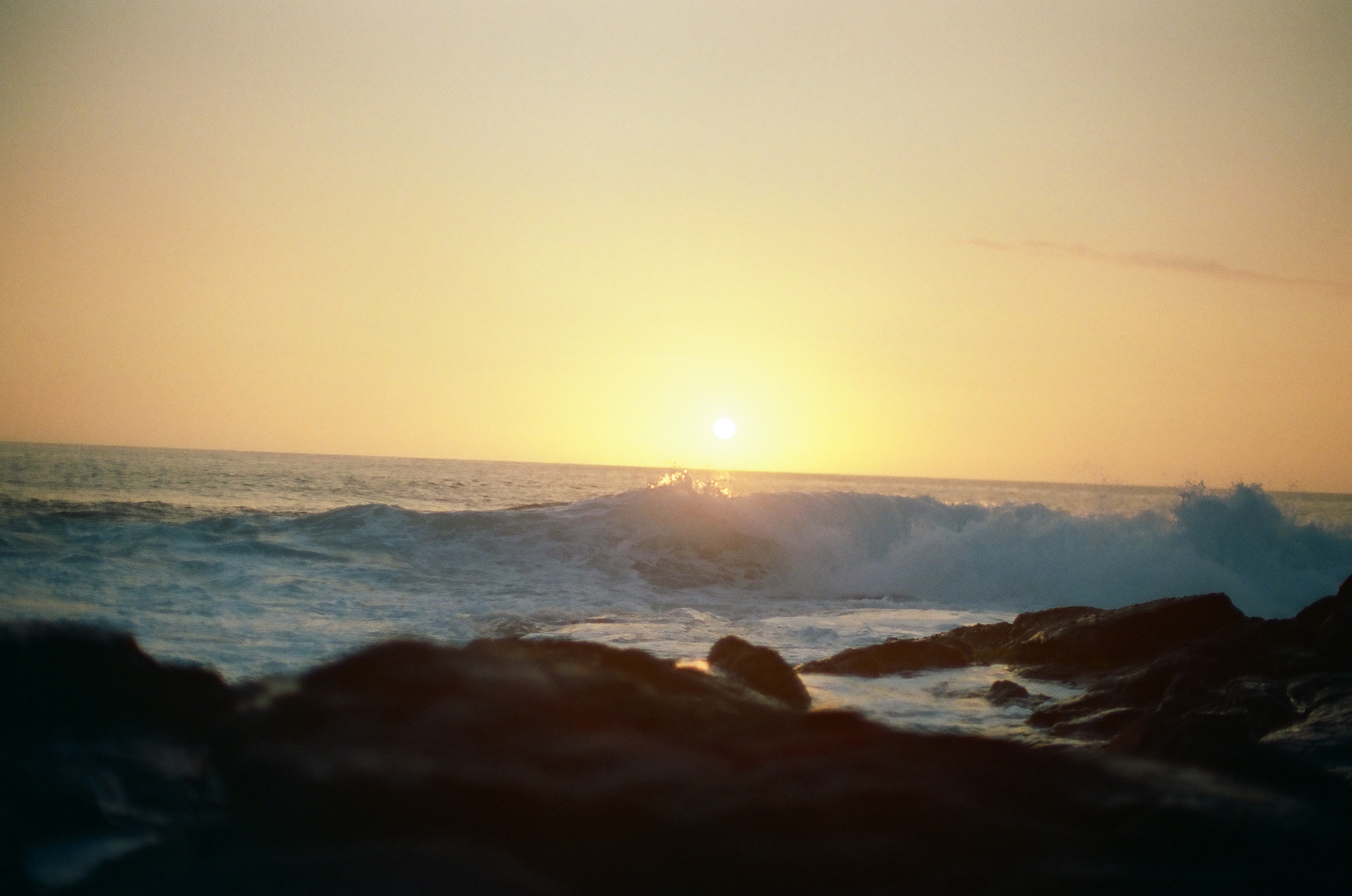 A broad wave collapses into white foam across black rocks as the sun nears the horizon.