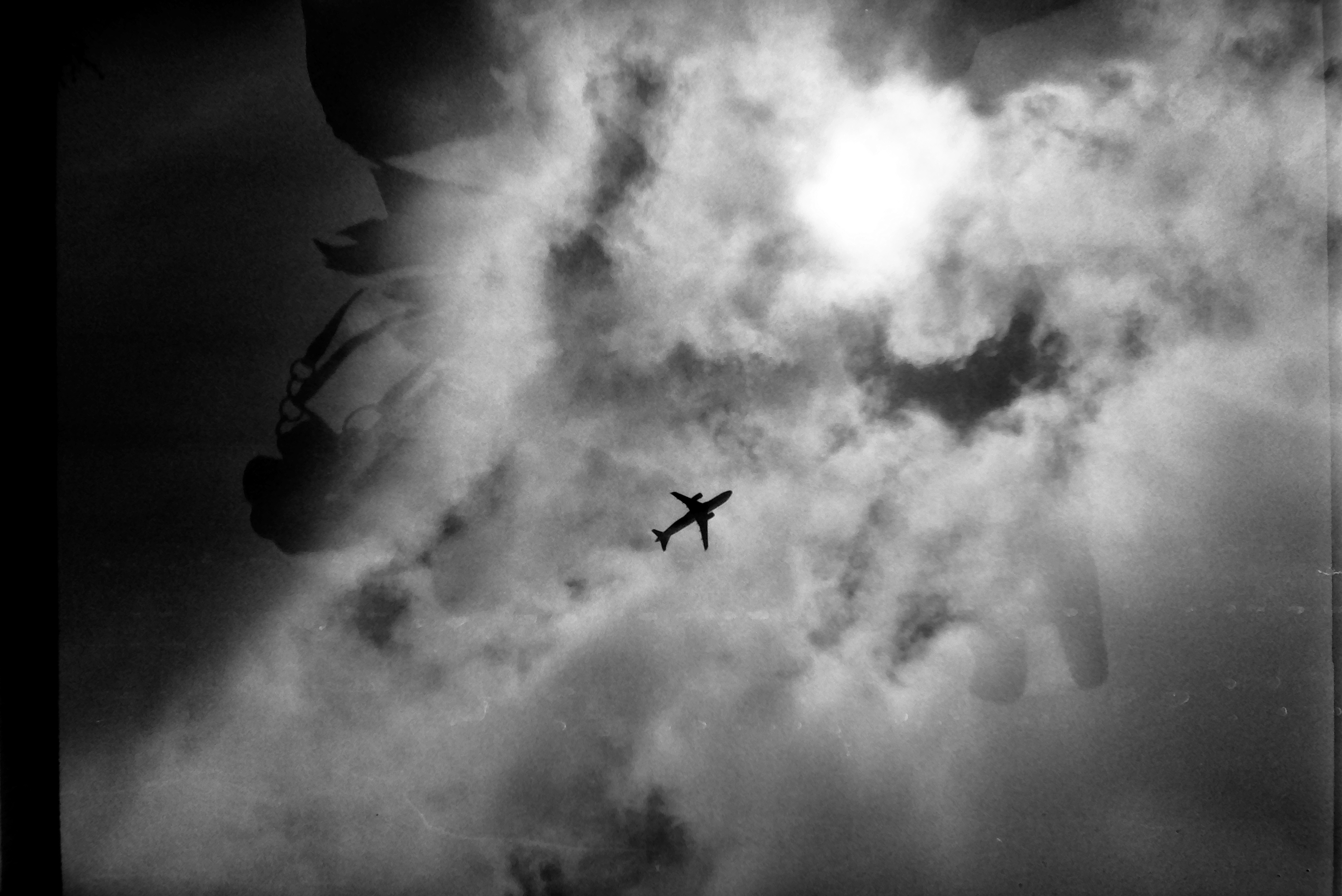 A small airplane silhouette crosses bright, broken cloud with the sun glaring near the top, and a faint darker shape overlays the left side.