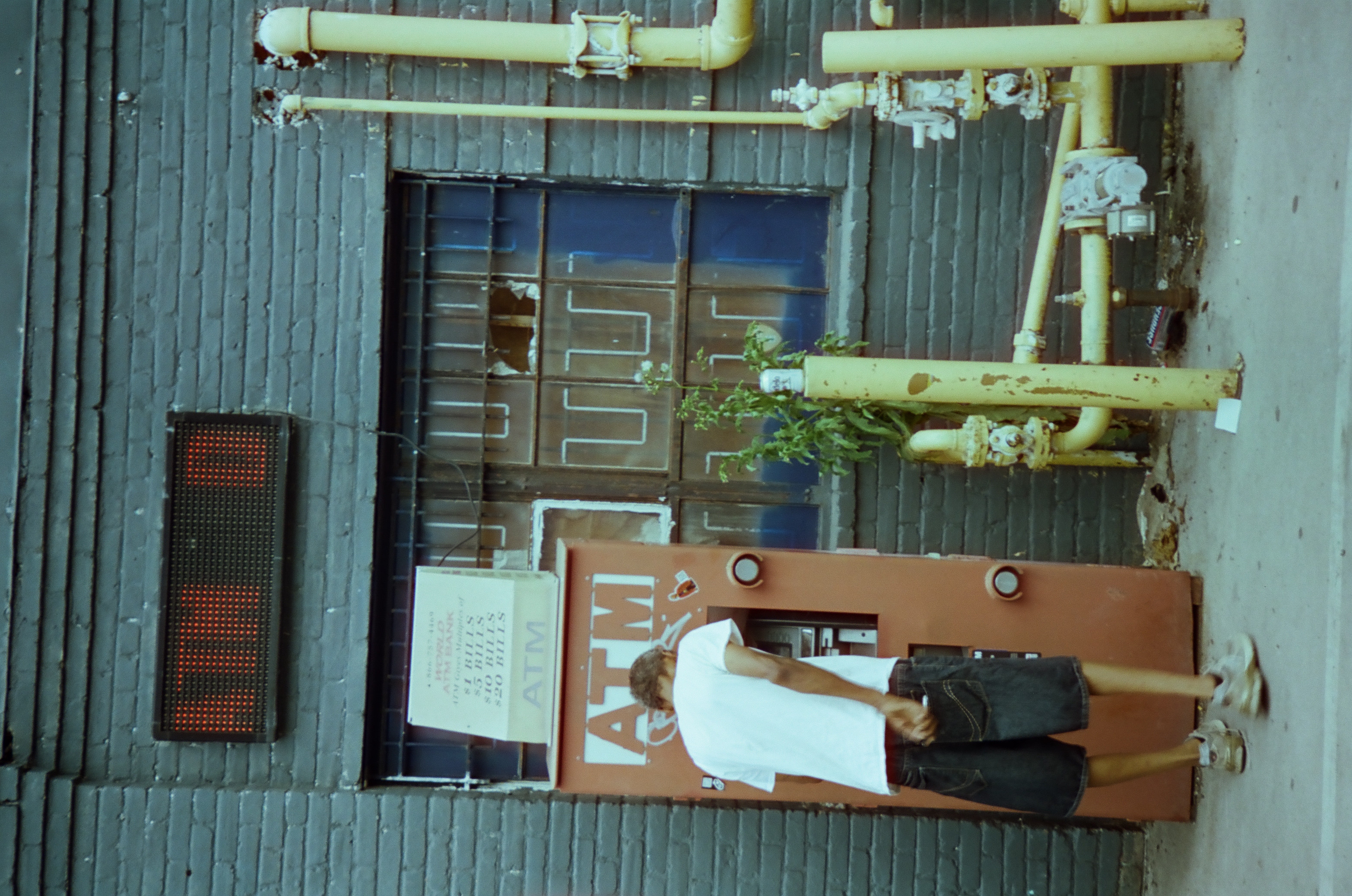 A person stands at a red ATM kiosk set into a gray painted wall beneath yellow gas pipes.