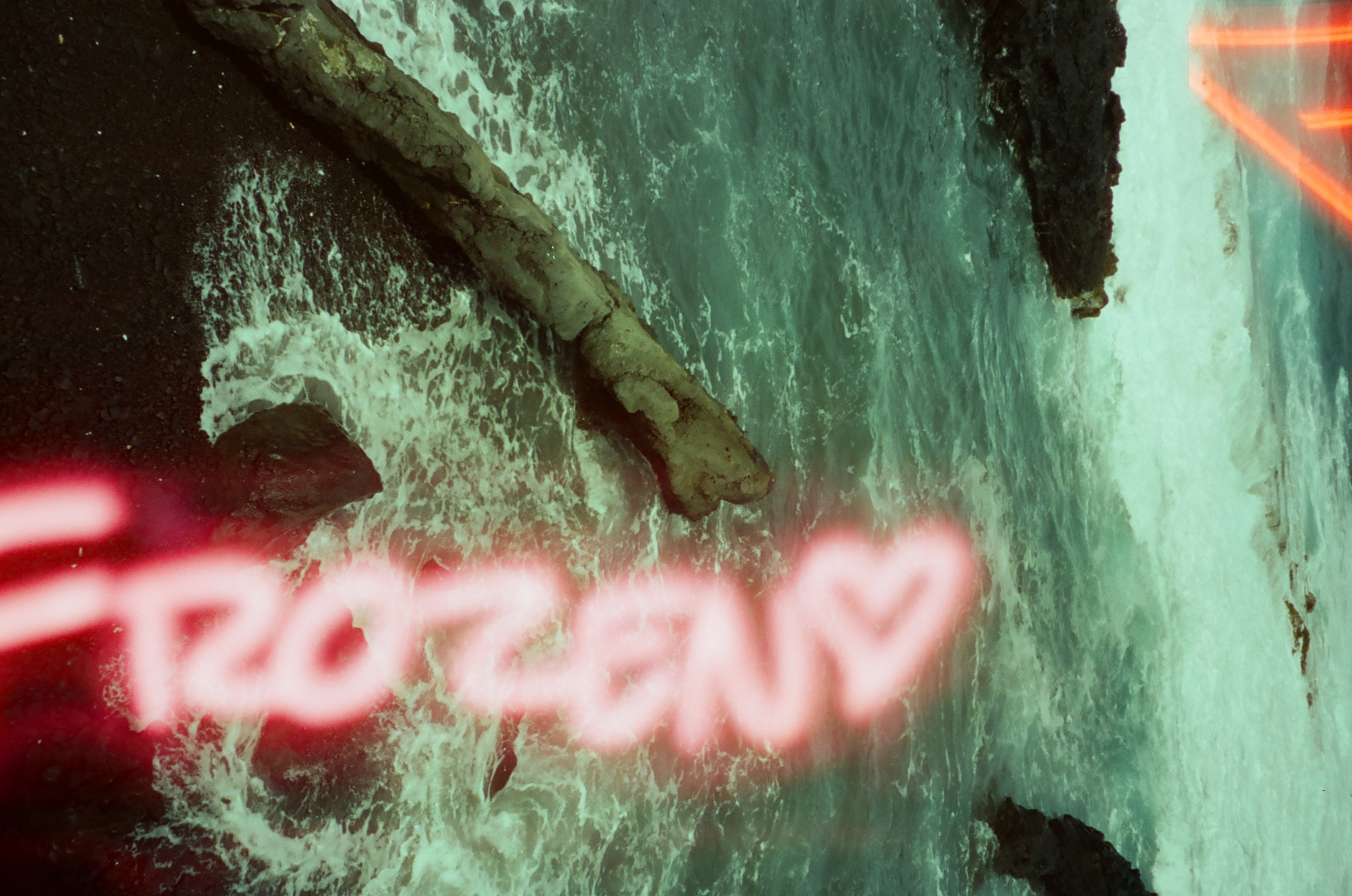 Foam-covered dark rocks and turquoise water fill the frame beneath glowing pink neon script that spells Frozen beside a small heart.