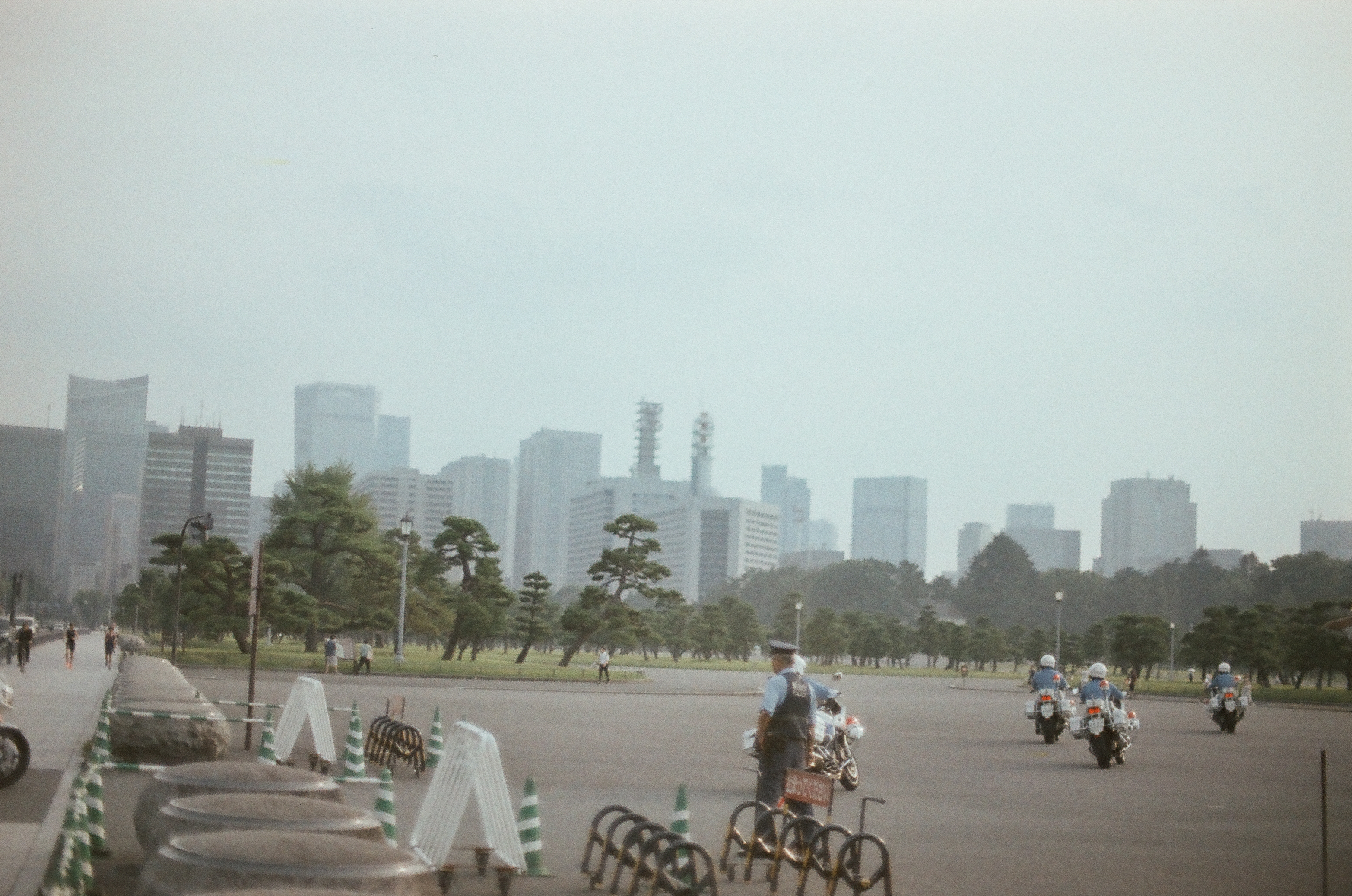 Uniformed riders on police motorcycles cross a broad paved plaza lined with striped traffic cones, beneath a hazy skyline of high-rise towers.