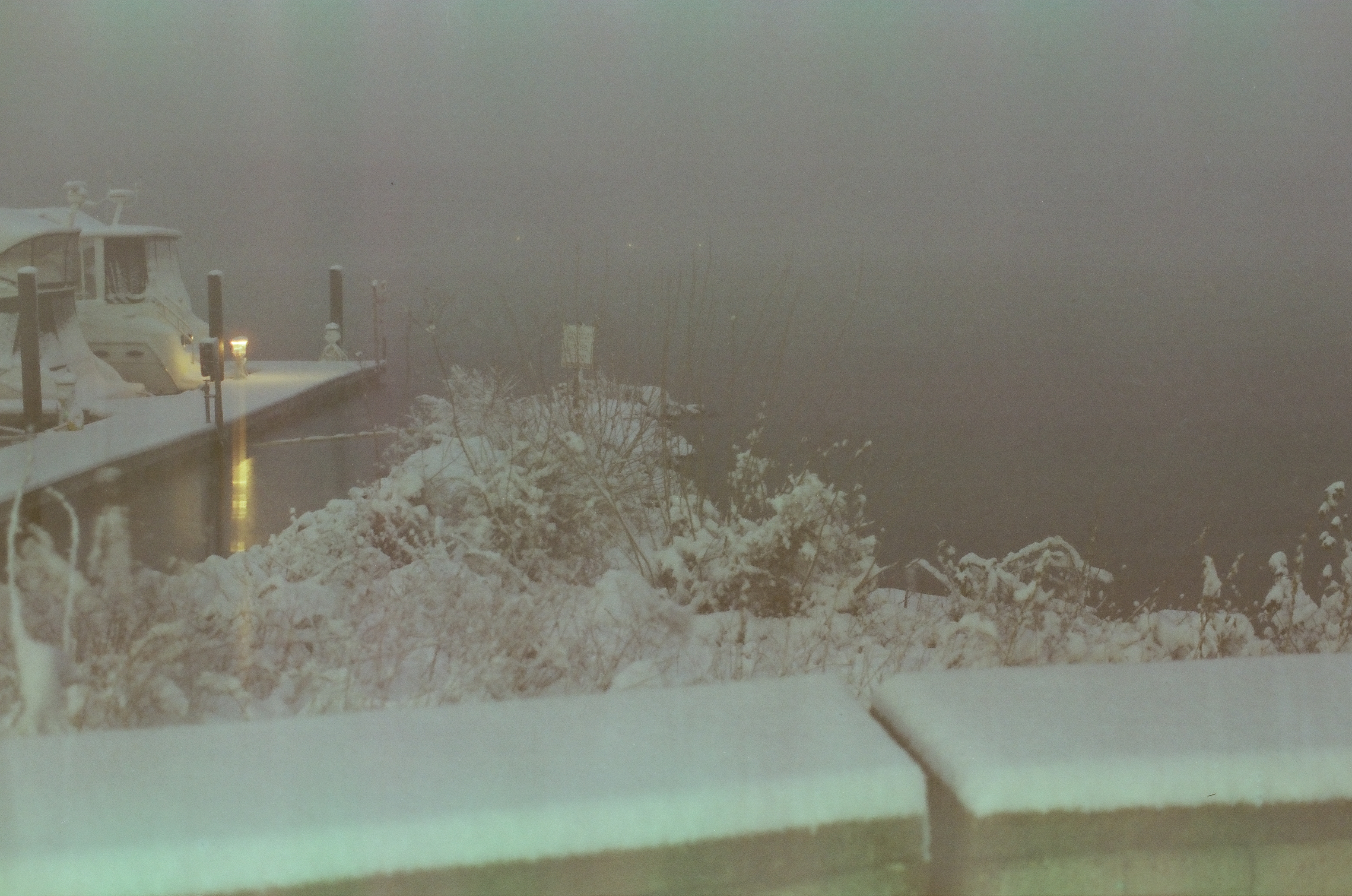 A snow-covered boat sits moored at a dock beside a glowing yellow light, its reflection stretching across dark water past snowy shrubs.