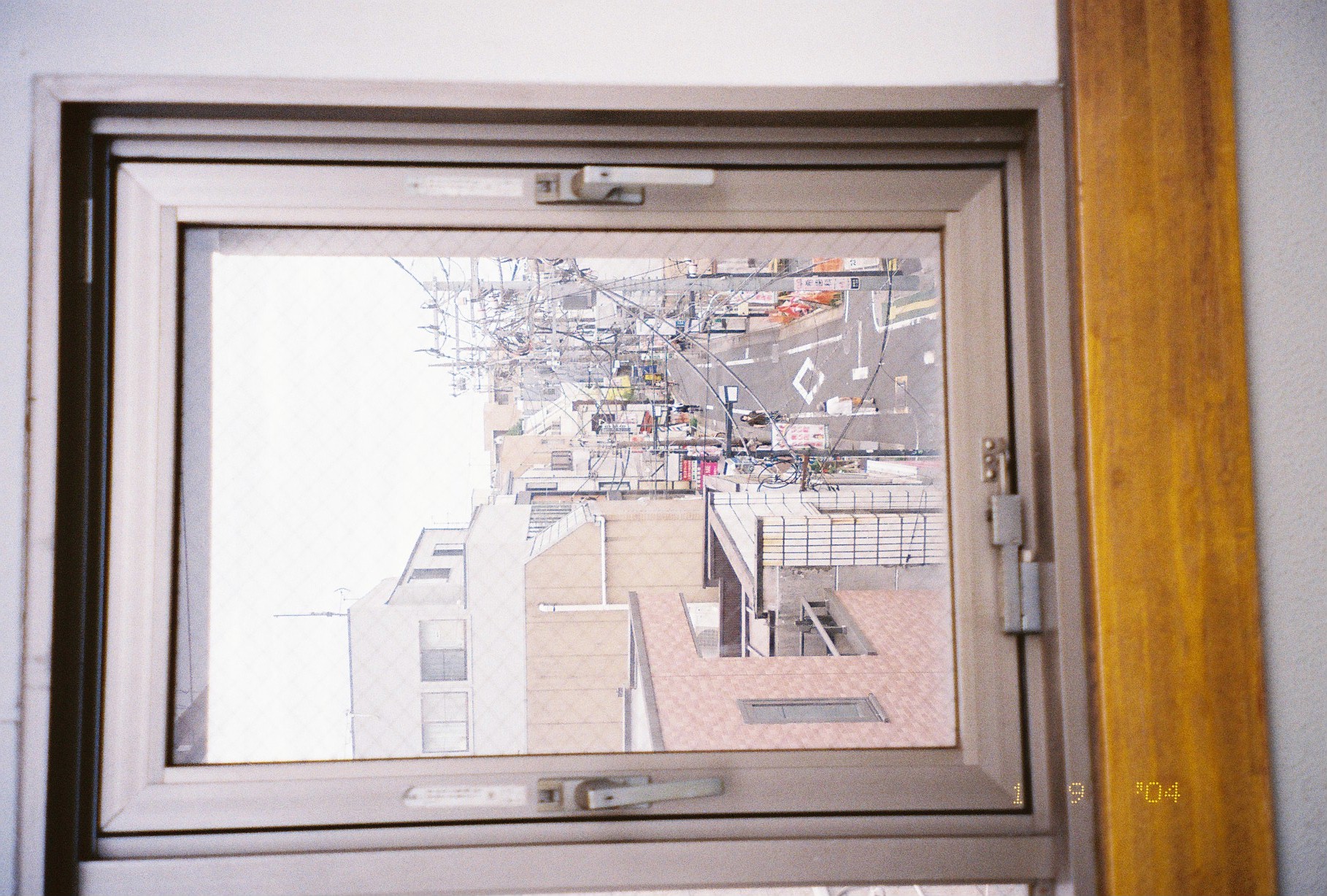 A window frame looks out over a narrow street lined with utility poles, tangled wires, and low buildings under white sky.