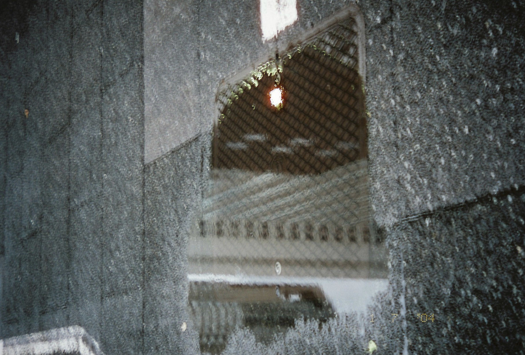 A puddle on dark paving reflects a tall latticed building and a dark grid with one small bright light spot, beside a white painted line.