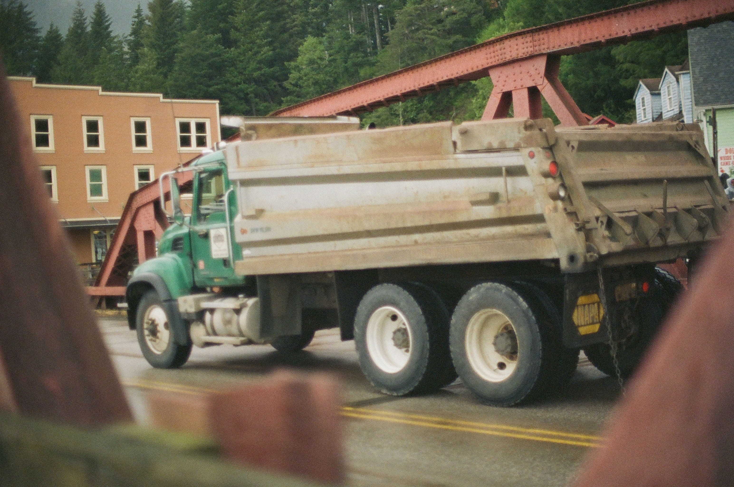 A green dump truck passes red industrial beams while blurred fingers partly close the foreground.