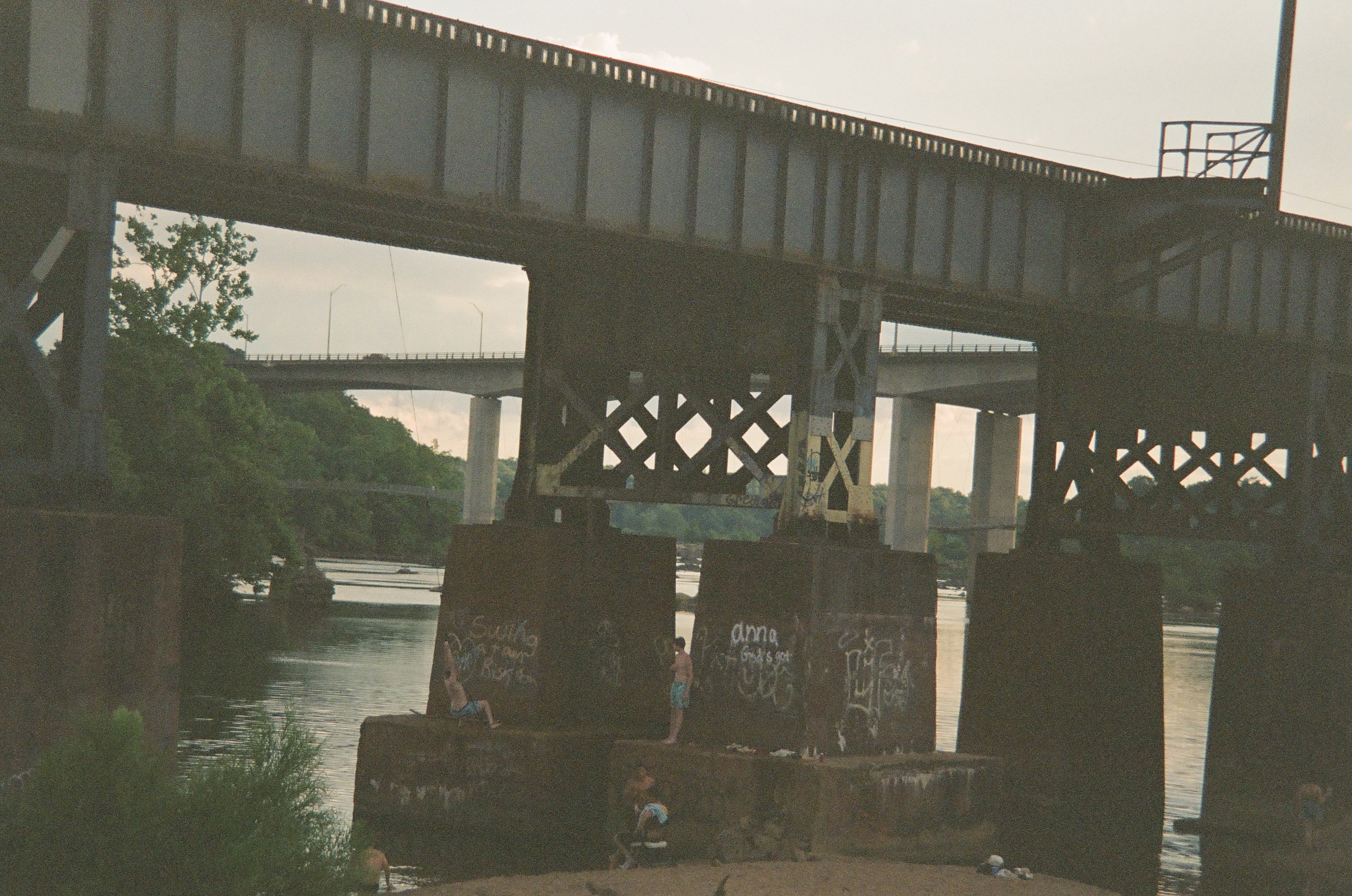 A steel railroad trestle and a highway bridge stack one above the other over the river, with two small figures on graffiti-covered piers below.