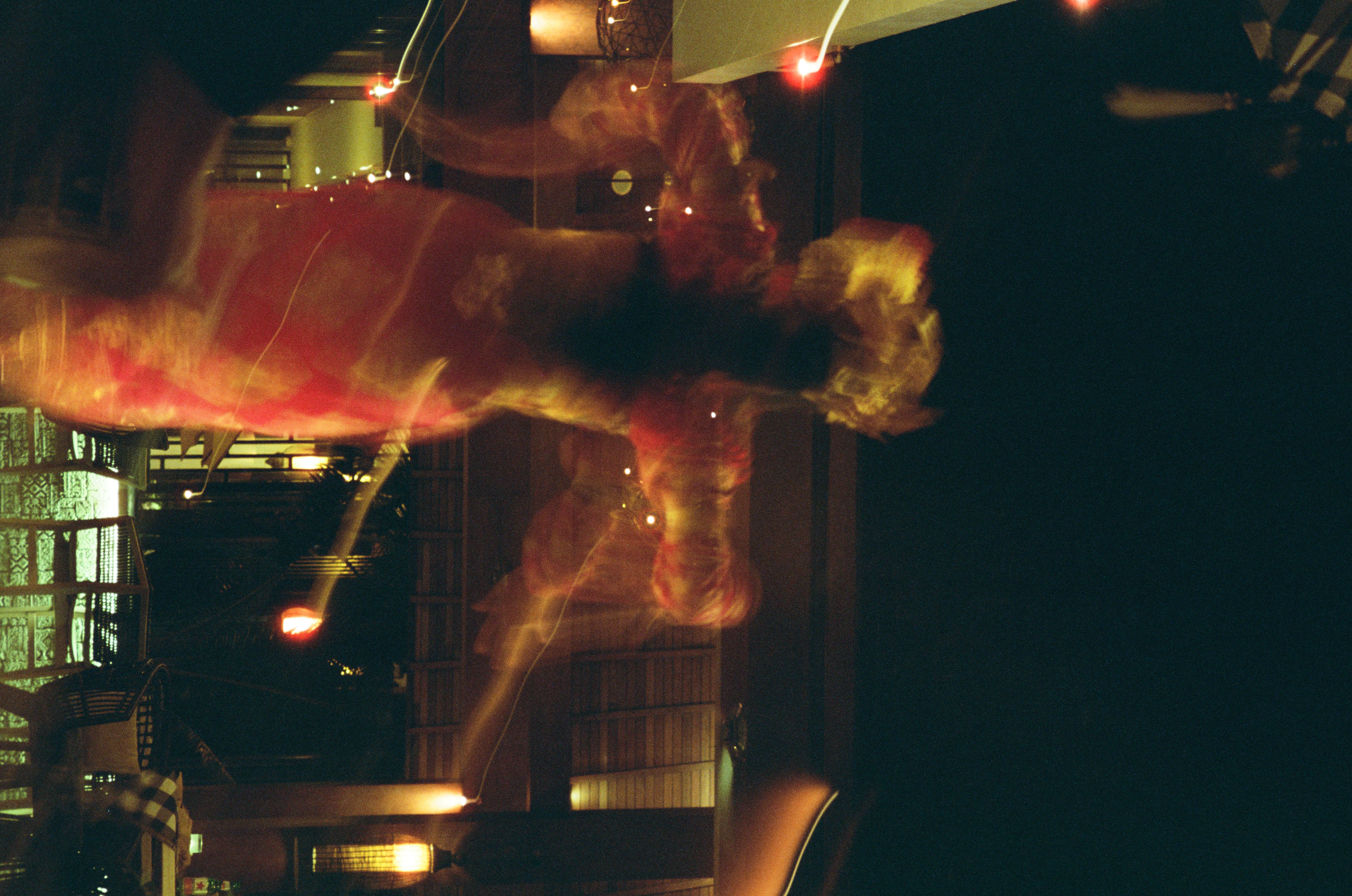 A warm red and gold blurred figure moves through multiple translucent layers beside strings of small lights and a patterned screen in near-total darkness.