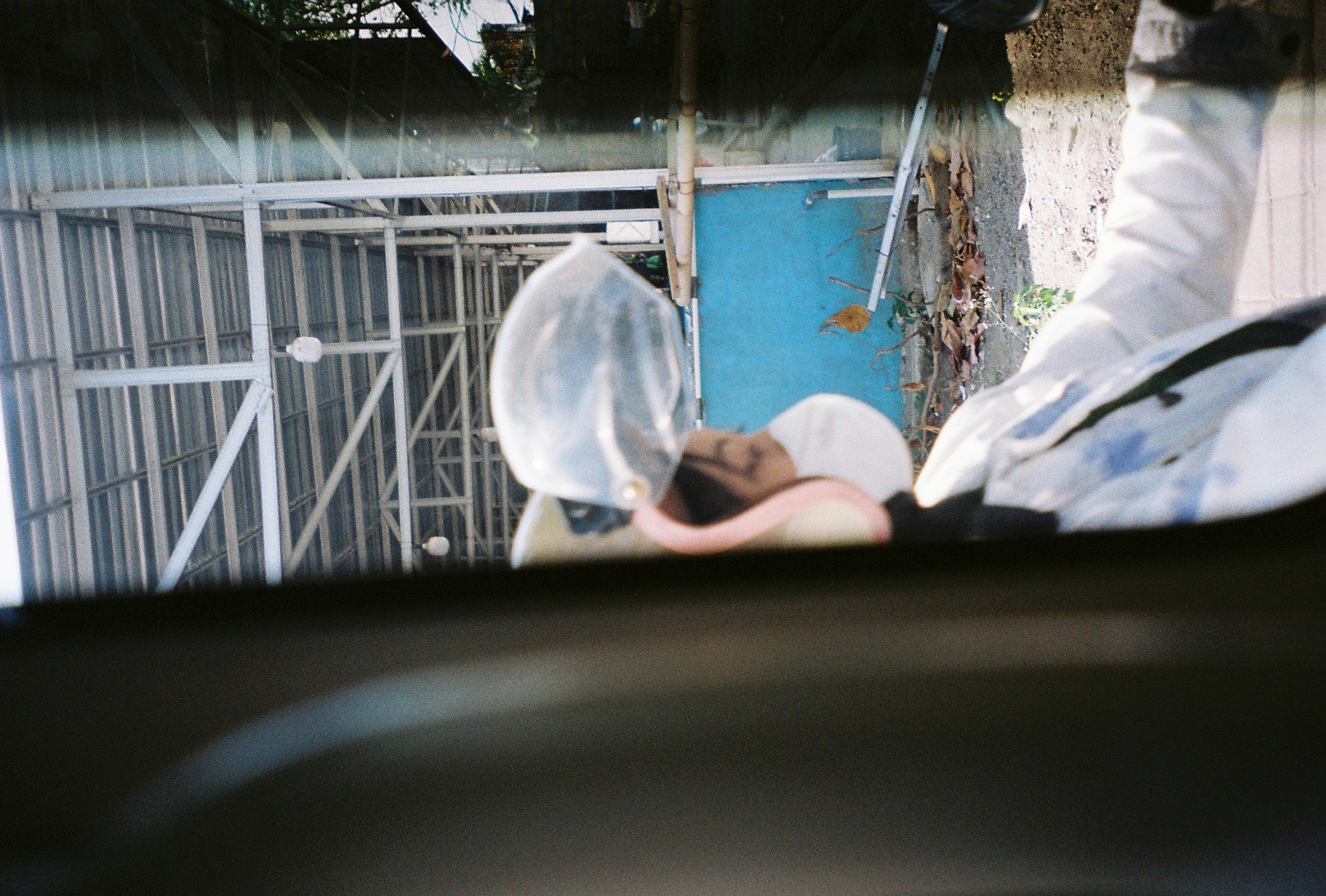 A masked, helmeted person in a pale jacket is partly visible between dark panels, with a blue wall and white roof framing behind.