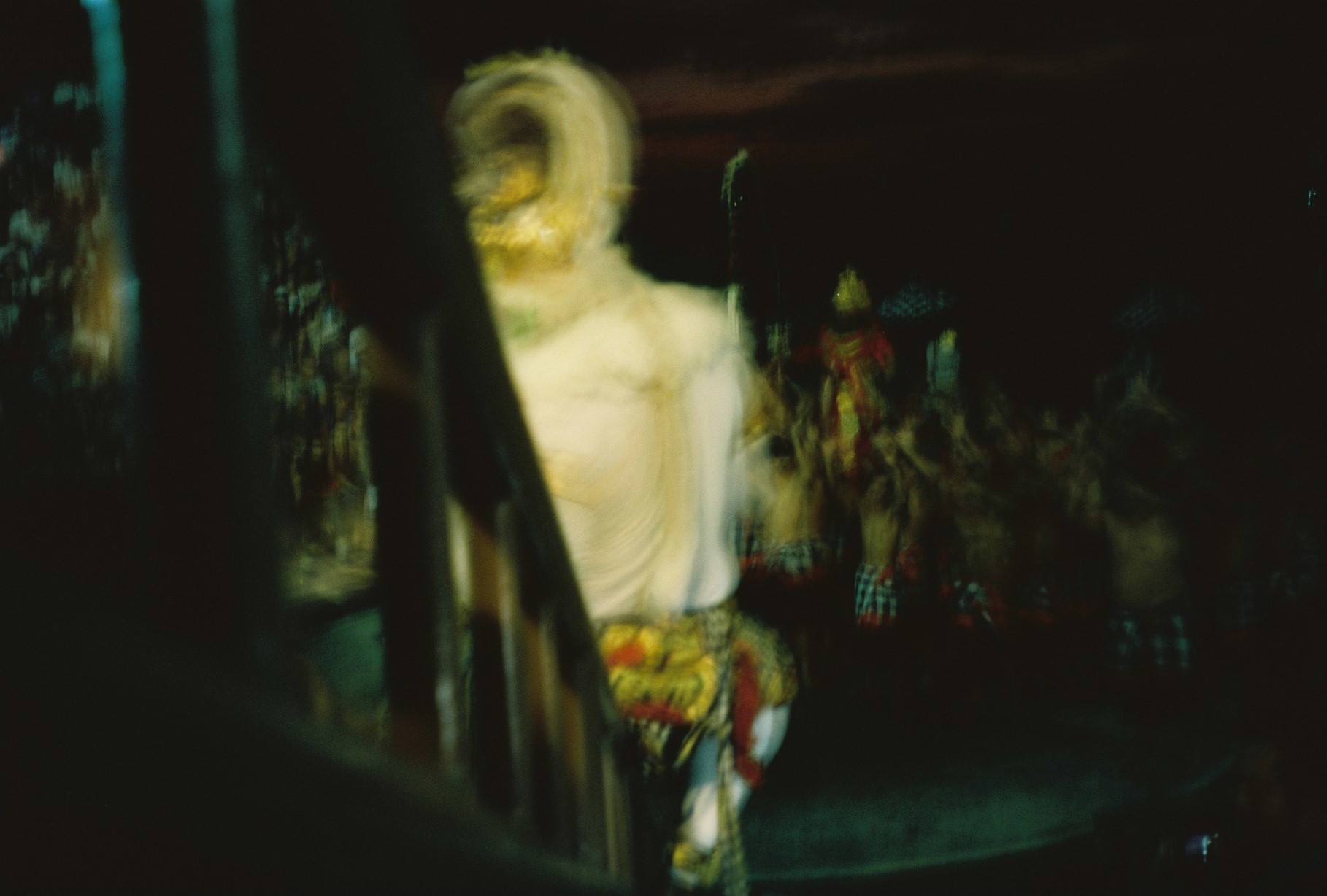 A pale costumed figure with a curled tail-like form stands near seated spectators in a dark crowded setting.