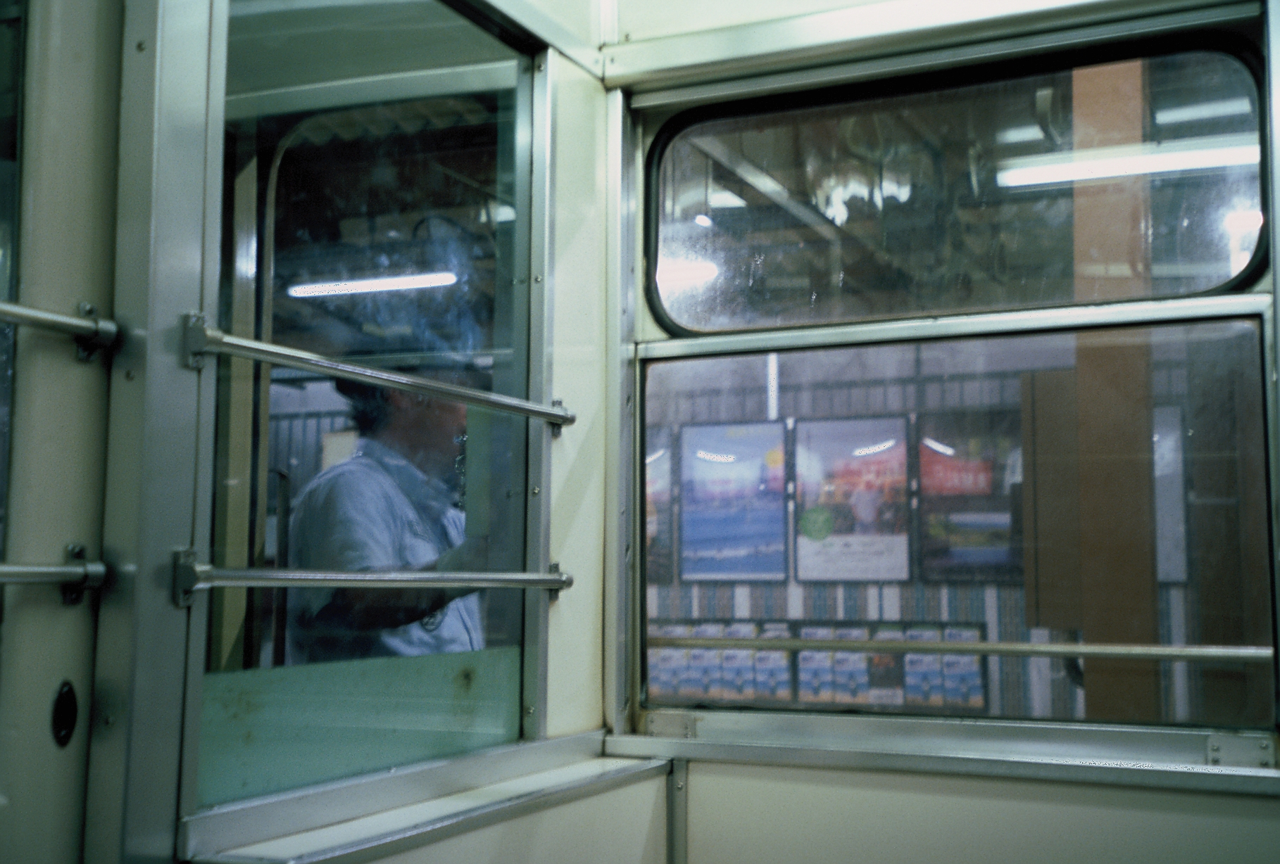 A passenger in a pale shirt is visible through one train car window, while framed posters show through the window beside it.
