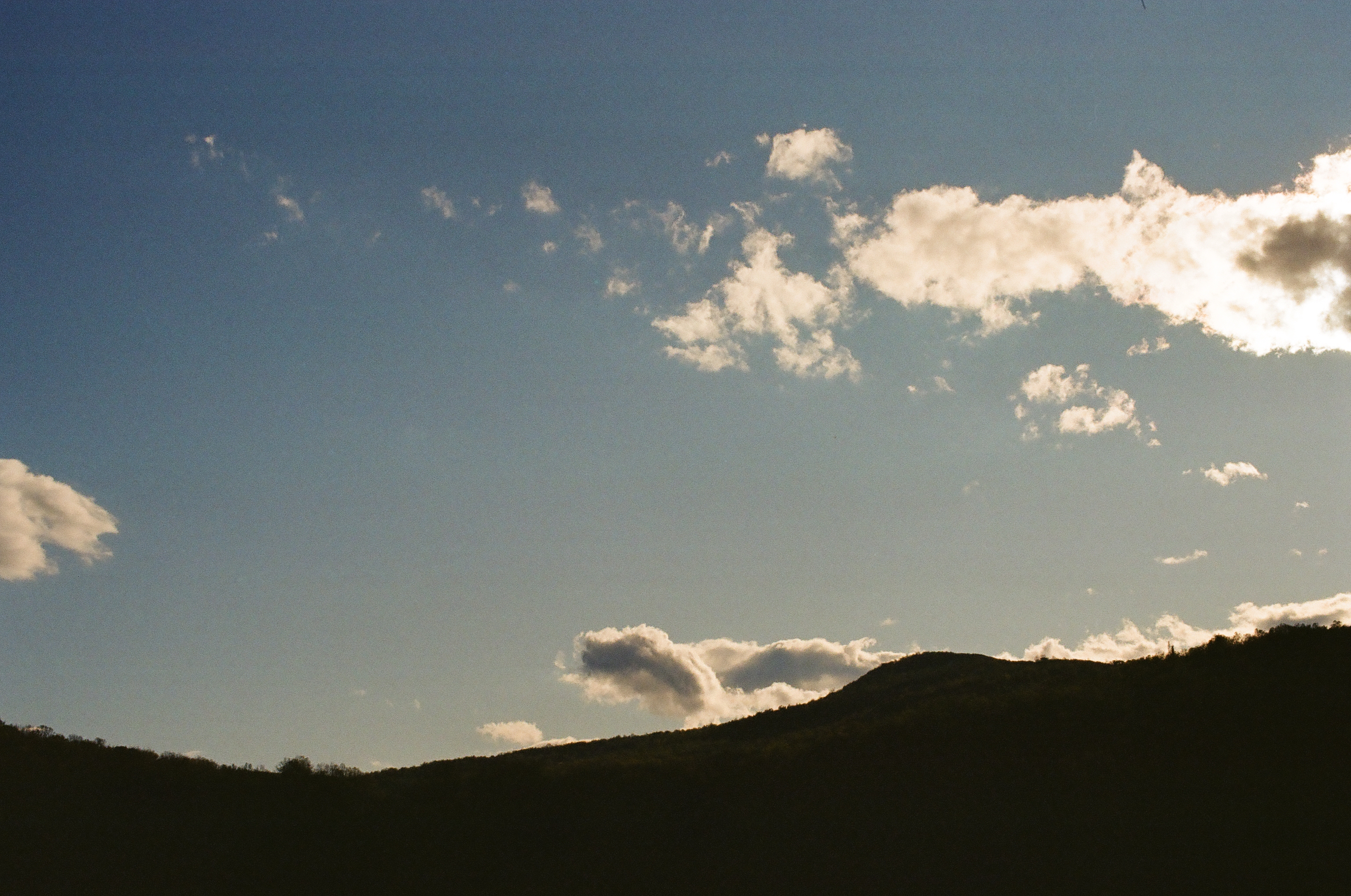 A hillside falls into full silhouette beneath a blue sky scattered with white clouds catching late-day light.