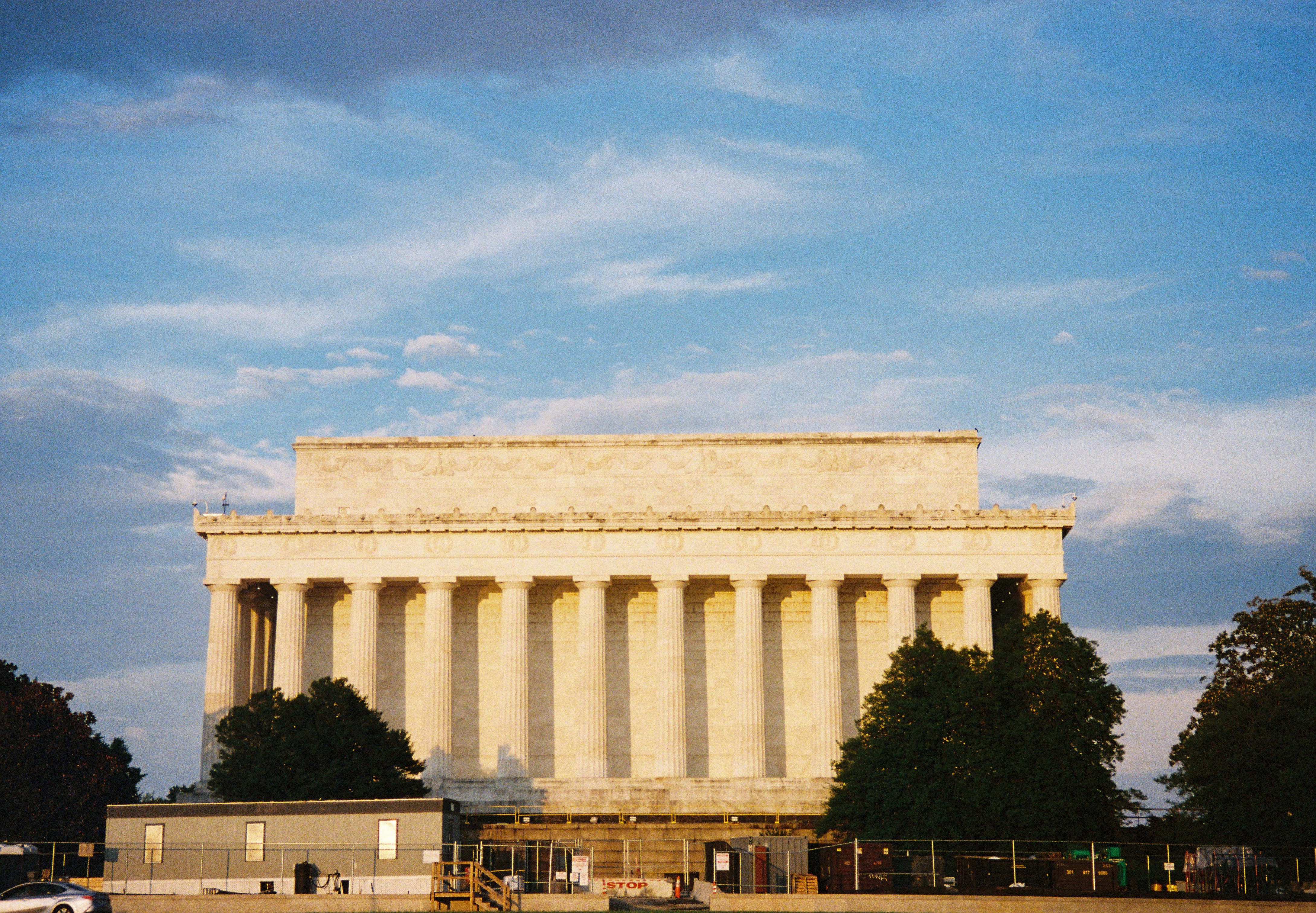 A broad pale memorial facade glows beyond dark trees, low barriers, and a deep blue sky.