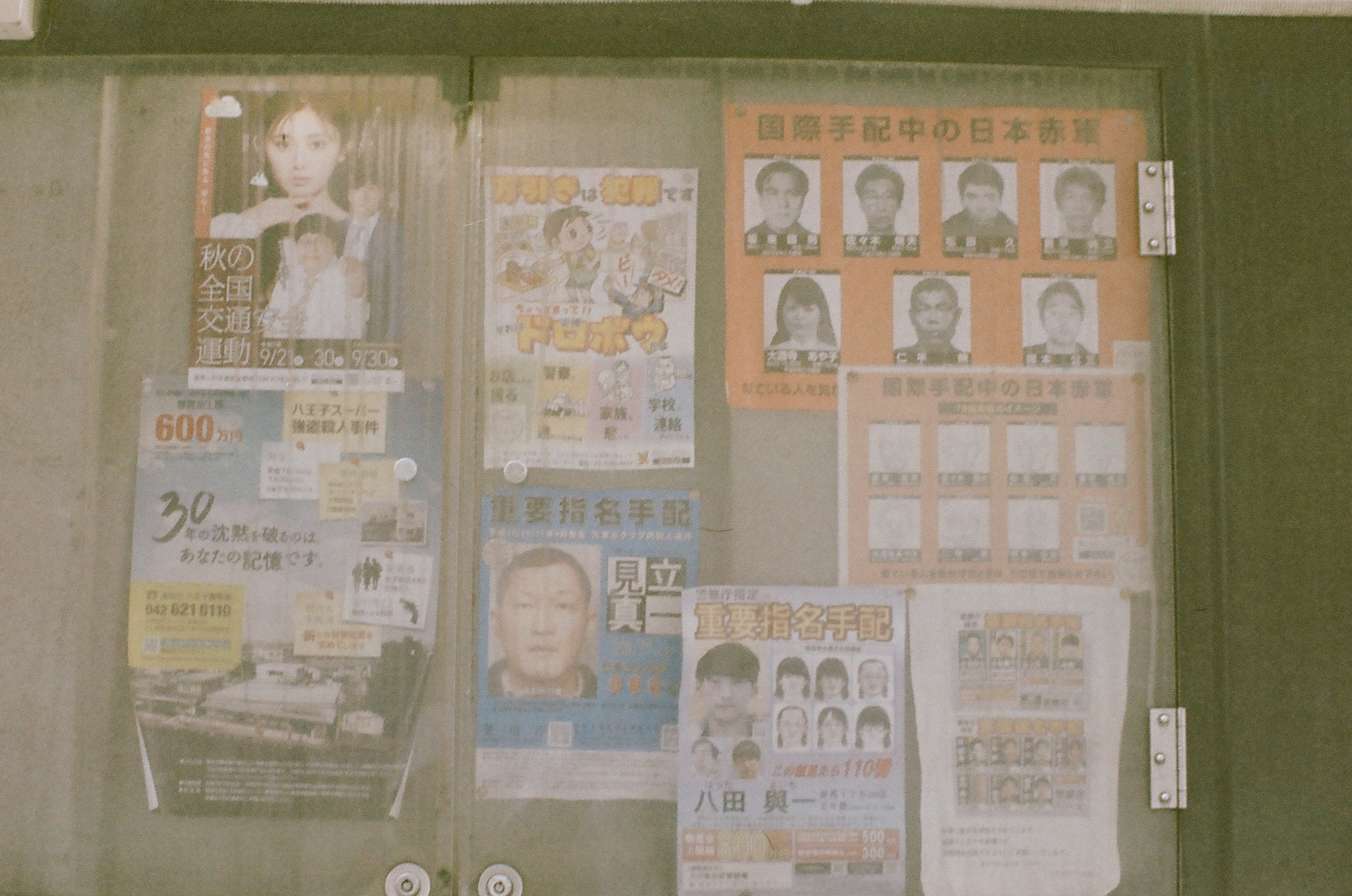 A public notice board behind glass displays layered posters, including grids of portrait photographs and a traffic-safety flyer.