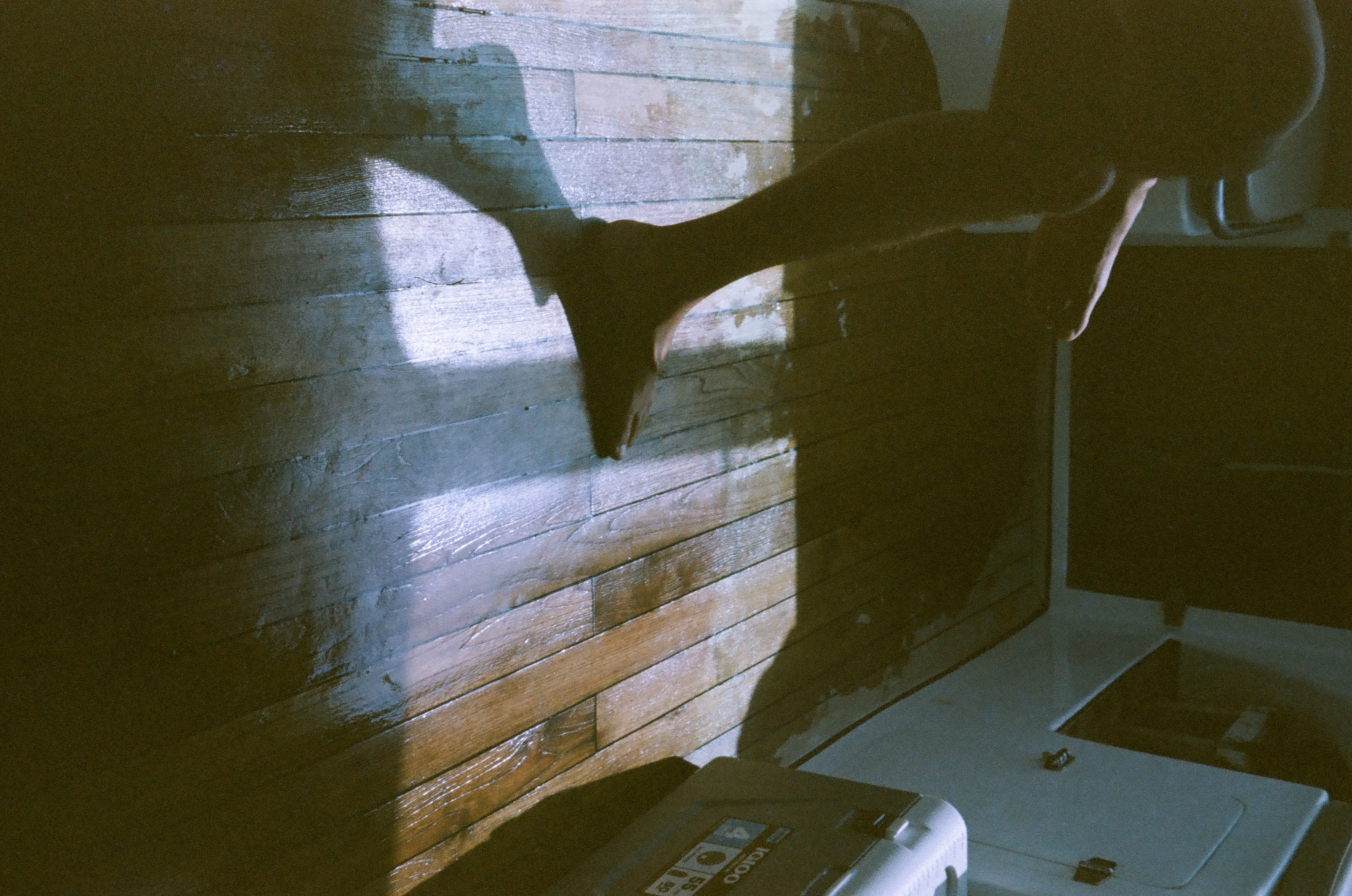 A seated person’s bare legs and feet cross a patch of sun on a wooden floor inside a dim boat cabin.