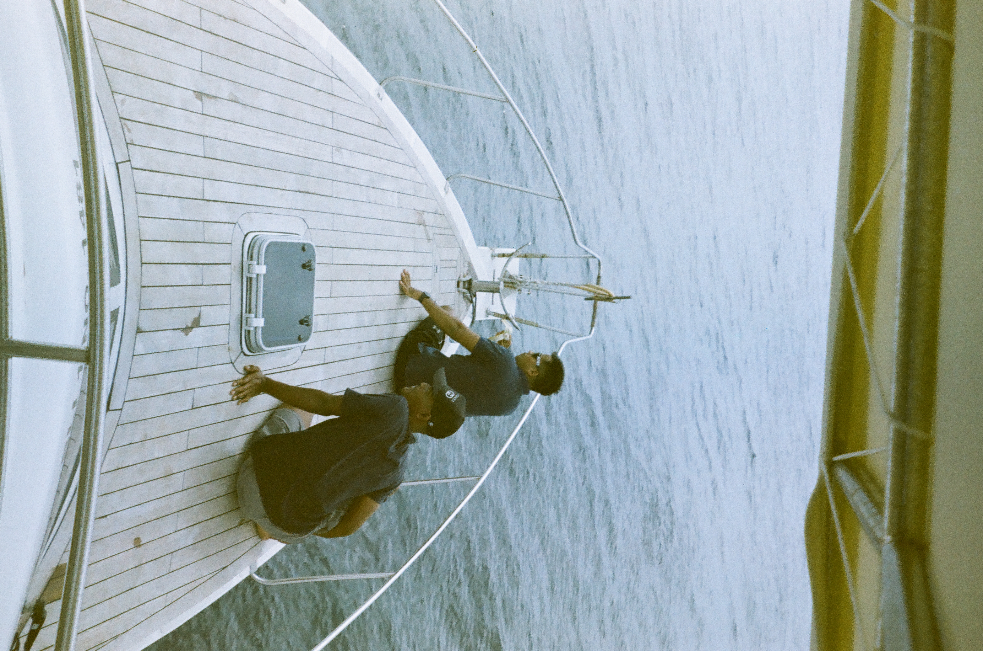 Two people sit on a pale plank bow facing the surrounding water beneath the dark edge of an overhead covering.
