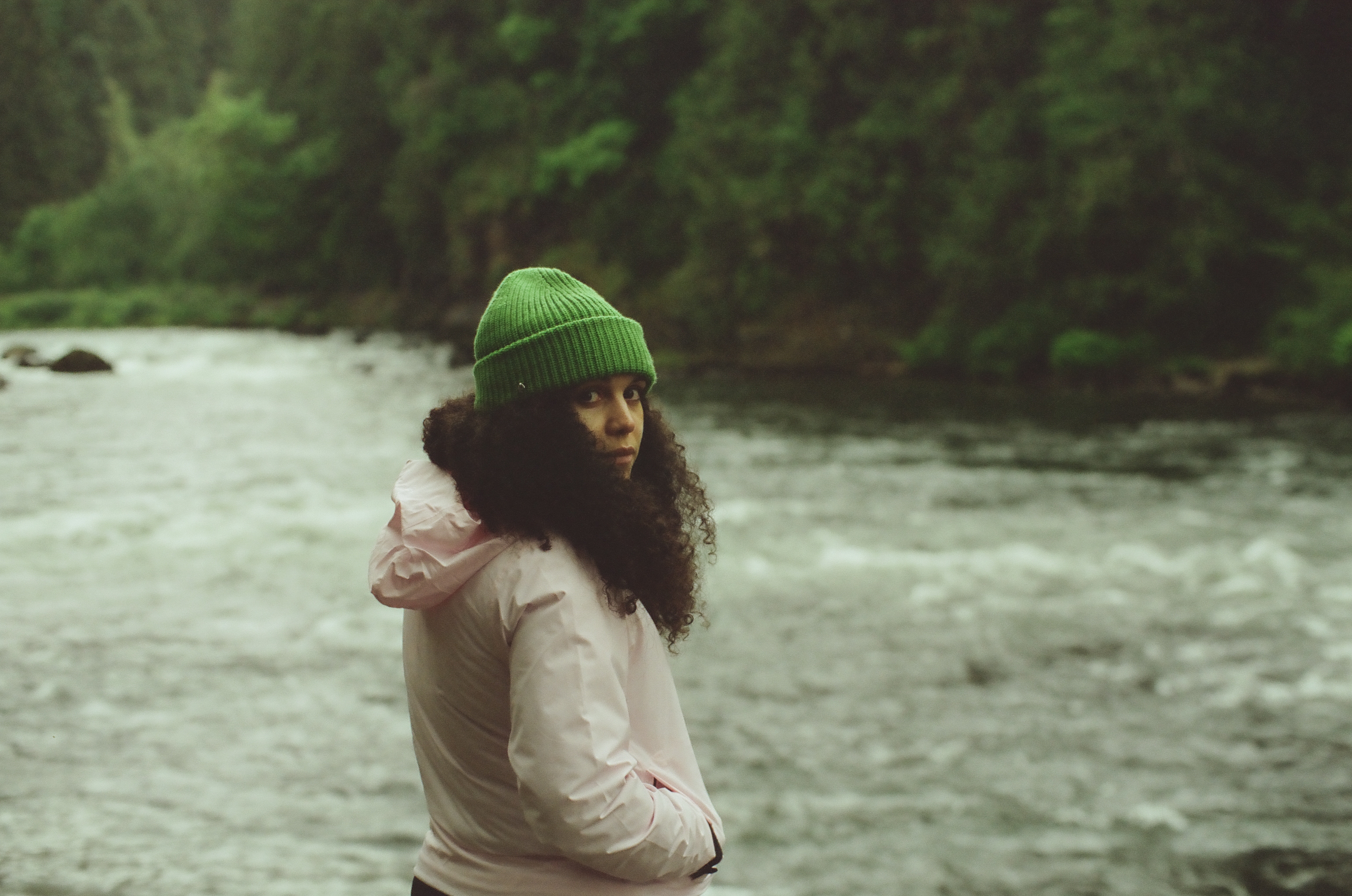 A person in a green knit cap and pale jacket stands beside a fast gray river.