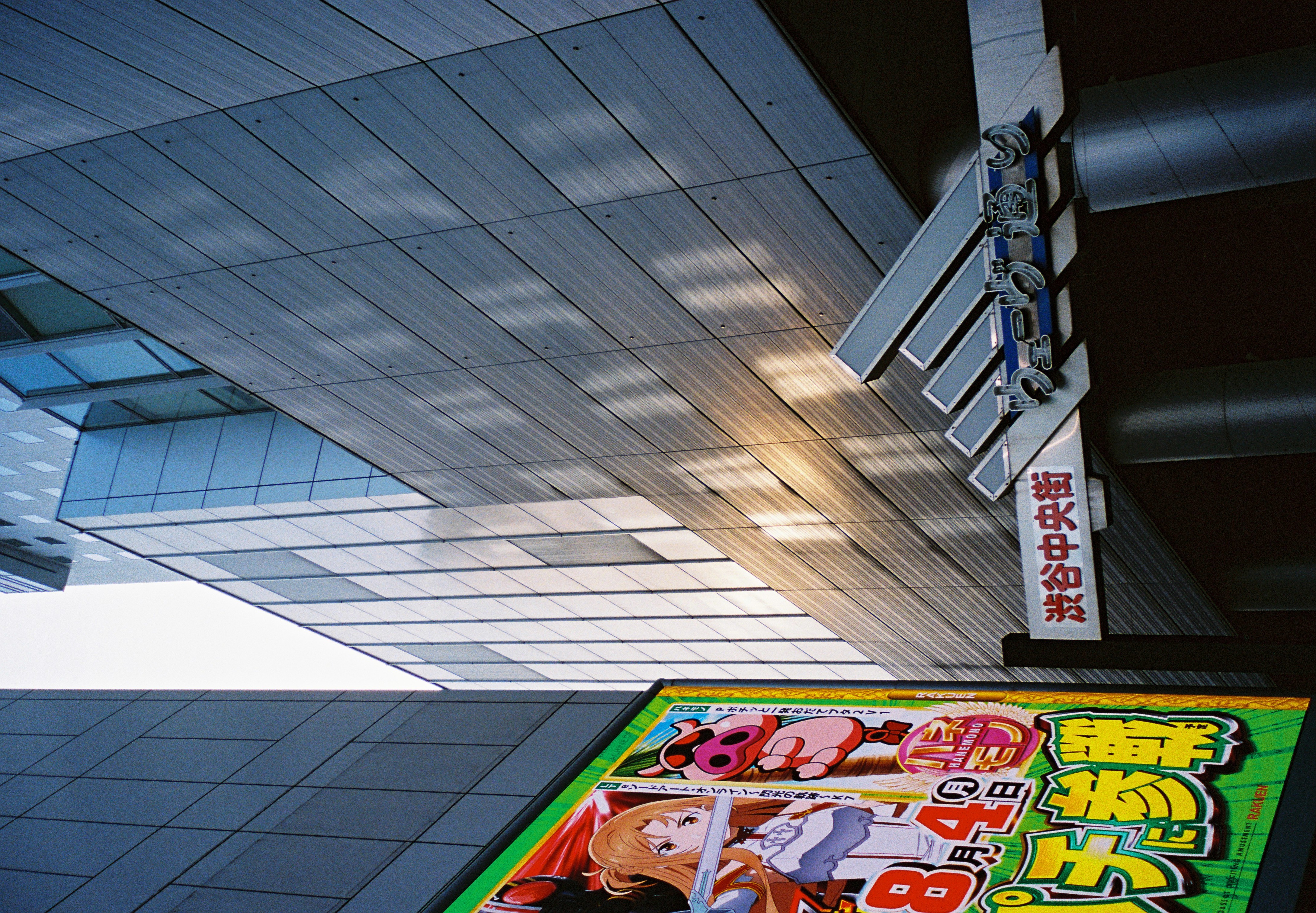 An illustrated anime-style billboard showing a sword-wielding character and a pink creature stands beside street signs reading '渋谷中央街' and 'ウェーブ通り.'
