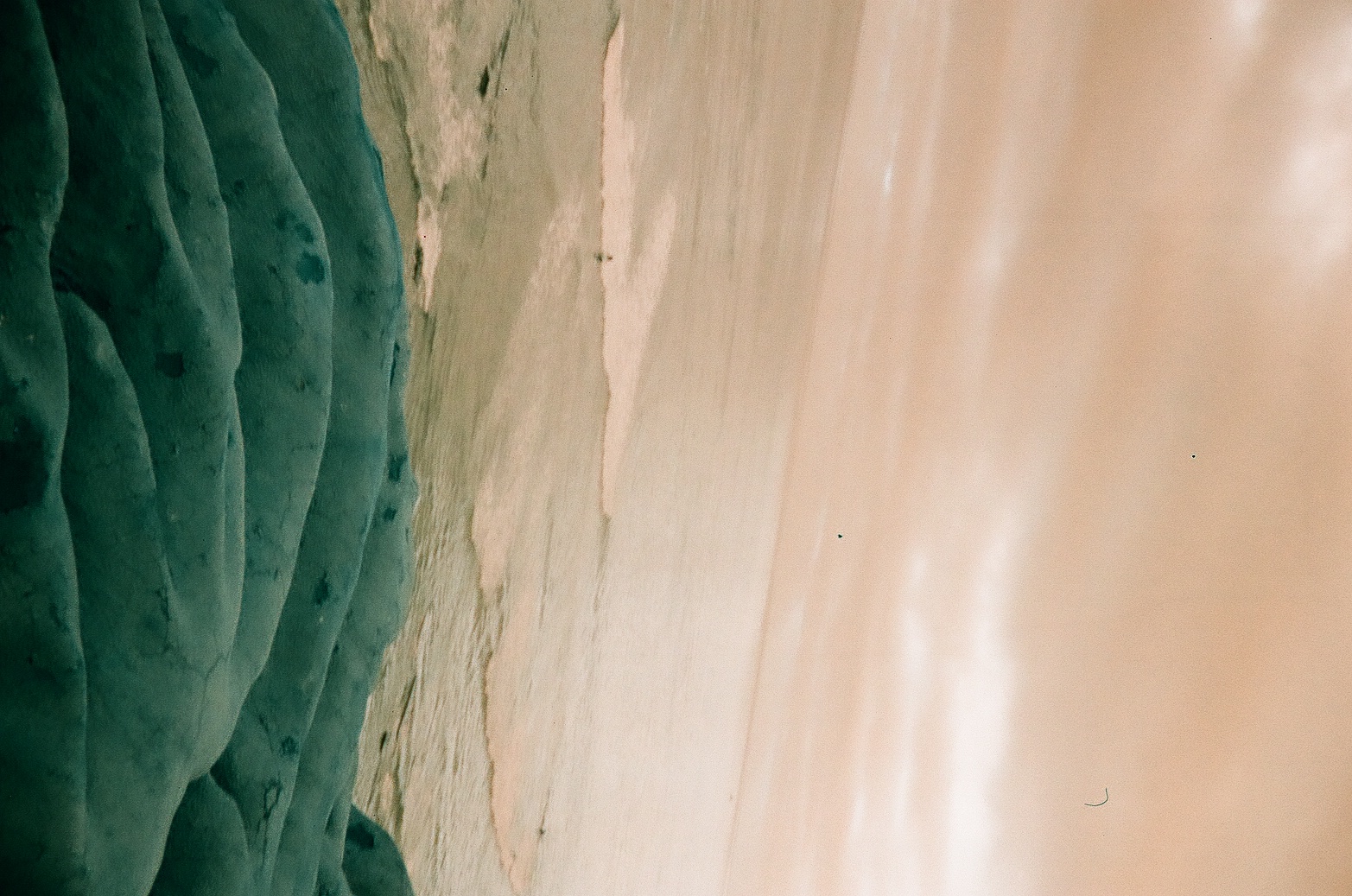 White ocean foam spreads over green-blue rocks beneath a pale peach horizon and softly clouded sky.