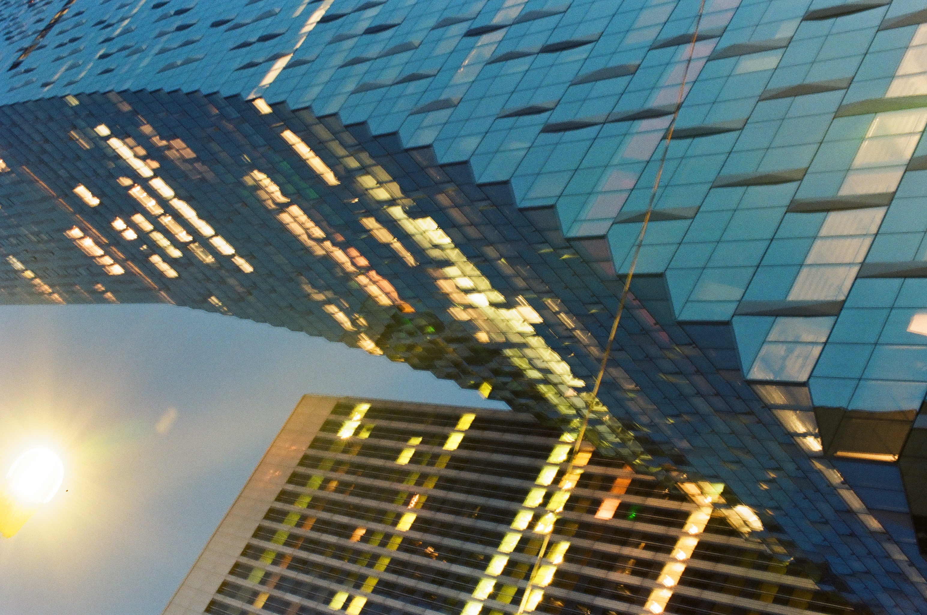 A blue glass tower rises between darker buildings beneath the flare of a streetlight.