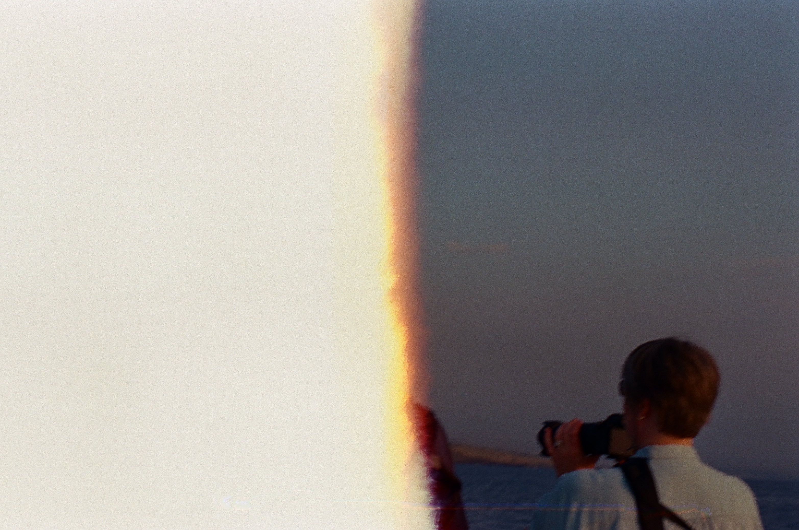 A vertical band of bright film flare divides pale sky from a cropped person holding a camera near the sea.