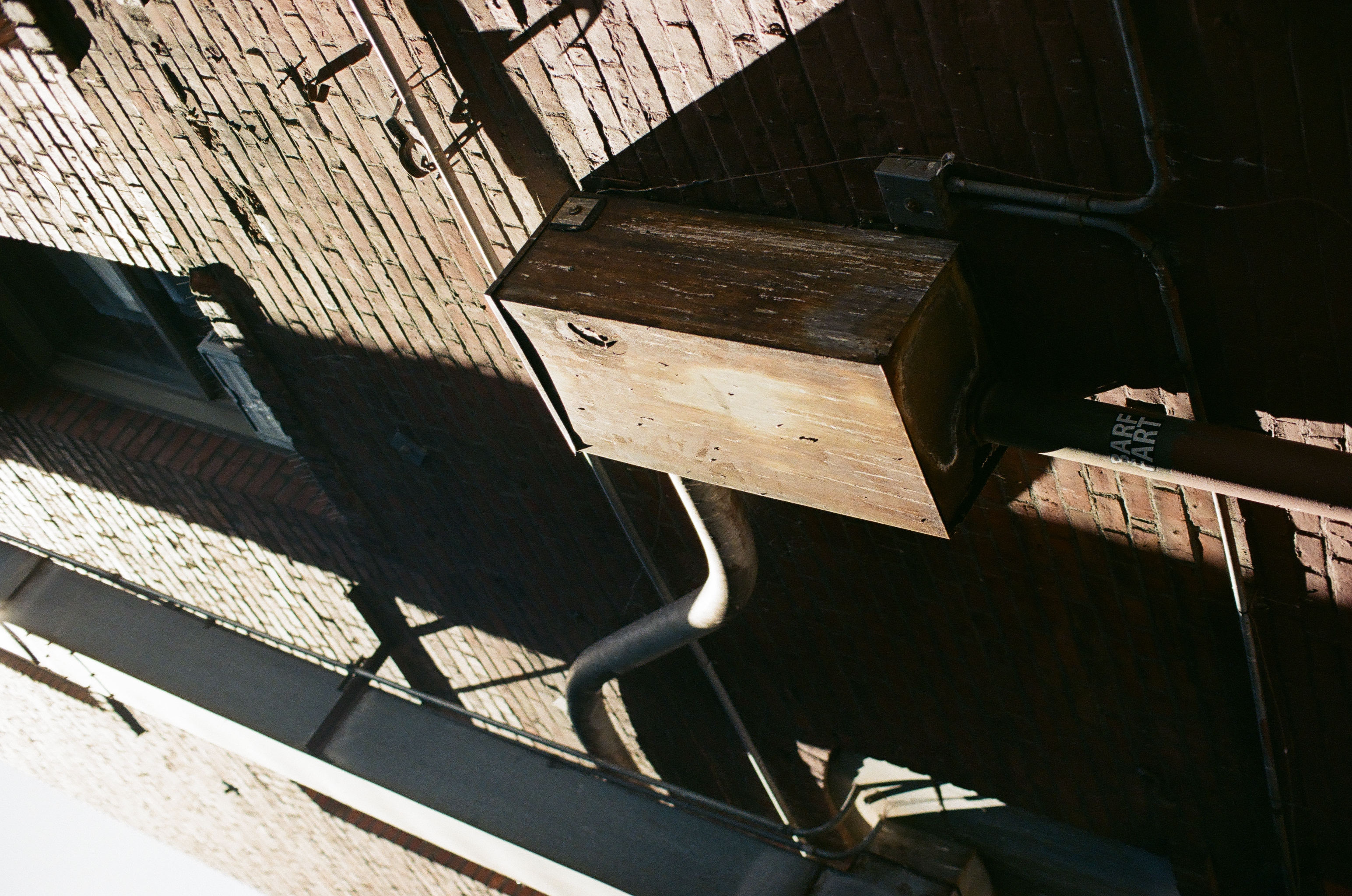 A weathered wooden utility box projects from a sunlit brick wall crossed by metal conduit.